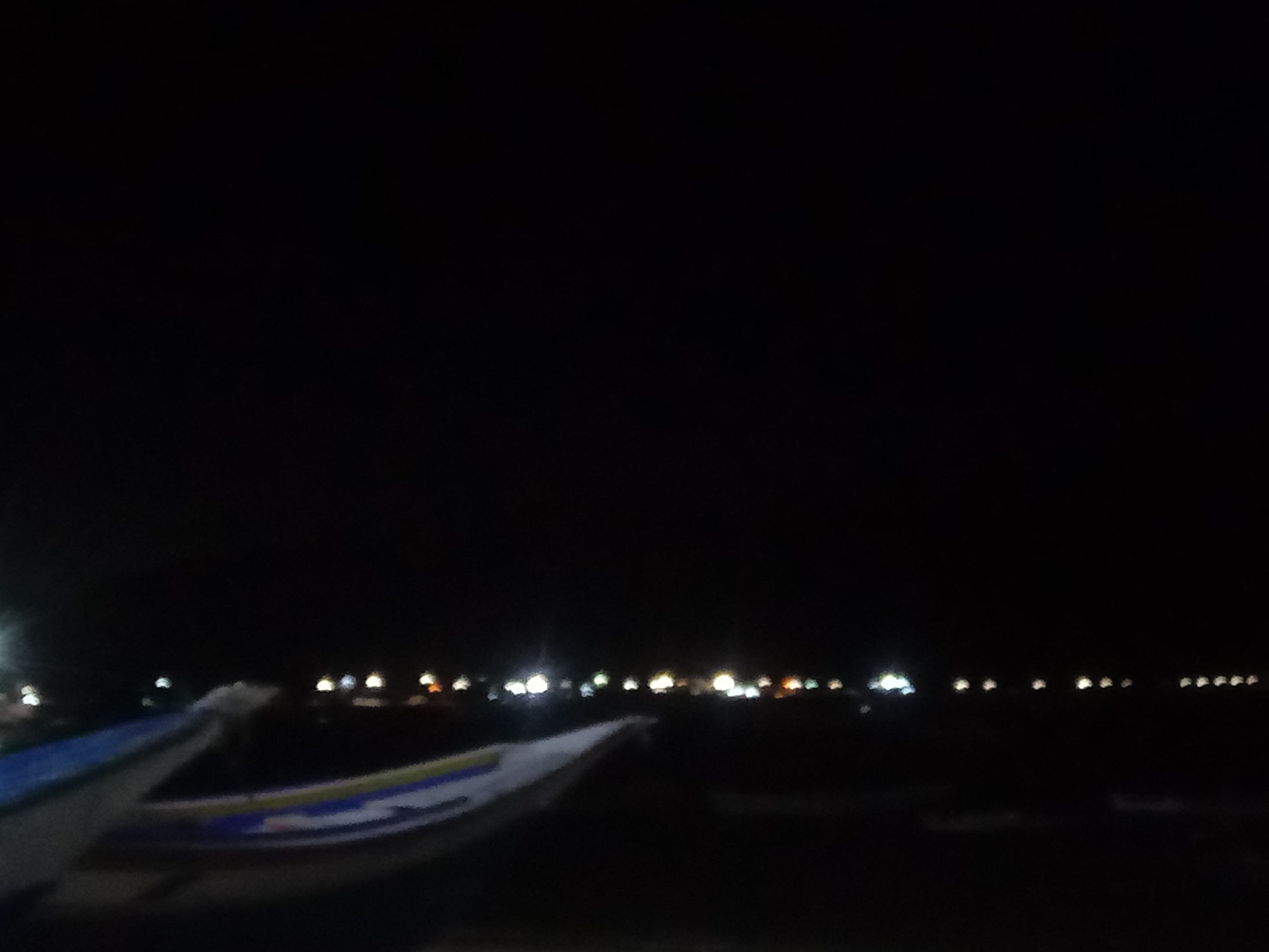 At night in Juan Griego, Venezuela, two boats sit close to the water. The horizon shows a series of lights, likely from buildings or streetlights in the distance. The sky above is dark.