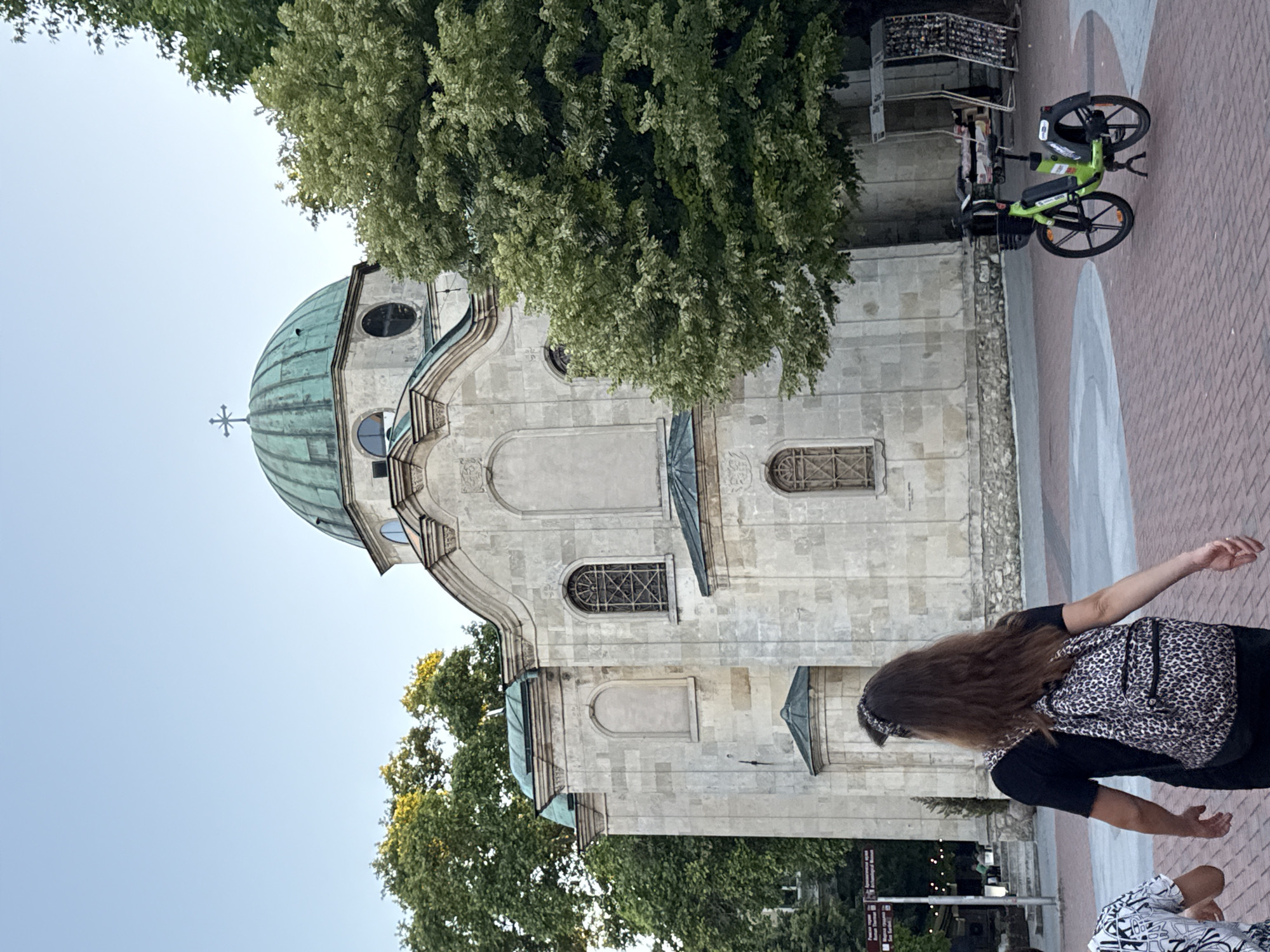 The image depicts a church with a green dome and a cross on top, built from light-colored stone blocks. The church has several arched windows with decorative metal grates. Adjacent to the church are lush green trees. In the foreground, a person with long brown hair, wearing a black t-shirt and a leopard-print backpack, walks away from the camera. To the left, another person is partially visible, wearing a patterned shirt. A bright green bicycle is parked on the brick-paved ground. The scene appears to be a public space, possibly a plaza, with some stalls or displays visible in the background. The sky is clear and pale blue.