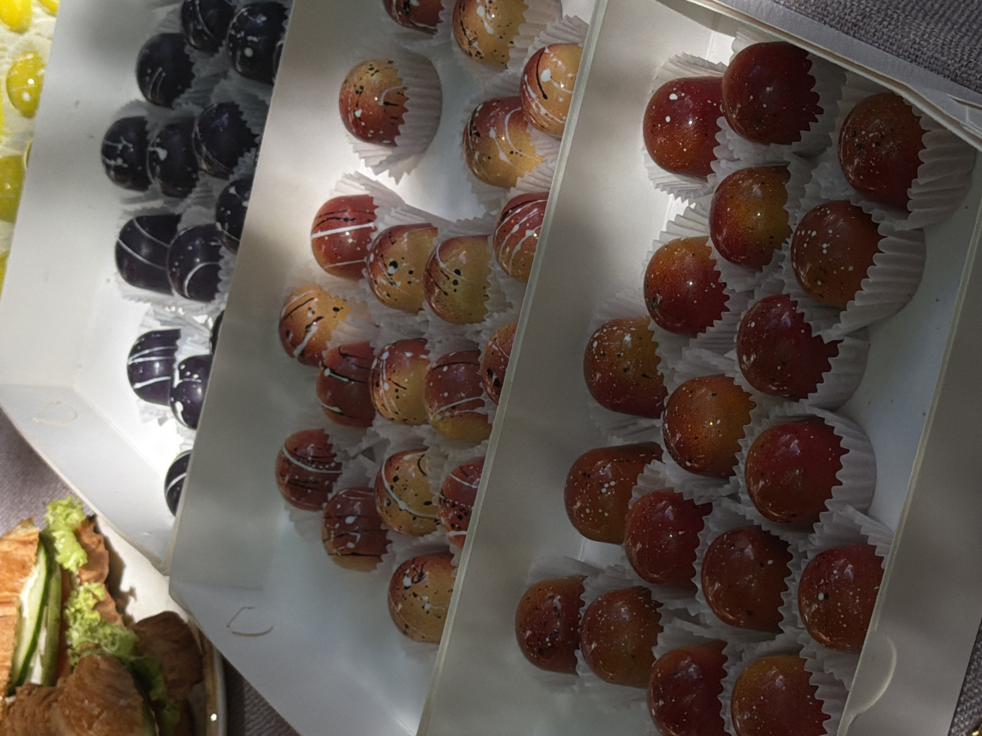The image shows three white boxes filled with gourmet chocolates. The box on the left contains dark purple chocolates with white accents, the middle box features orange and red chocolates with white details, and the box on the right contains predominantly red chocolates with speckled accents. All chocolates are individually placed in white paper wrappers. A partially visible plate of sandwiches is located in the bottom left corner of the image. The location is likely a confectionery or shop in Bishkek City, Kyrgyzstan. The scene depicts an array of decorative chocolates, potentially for sale or display.