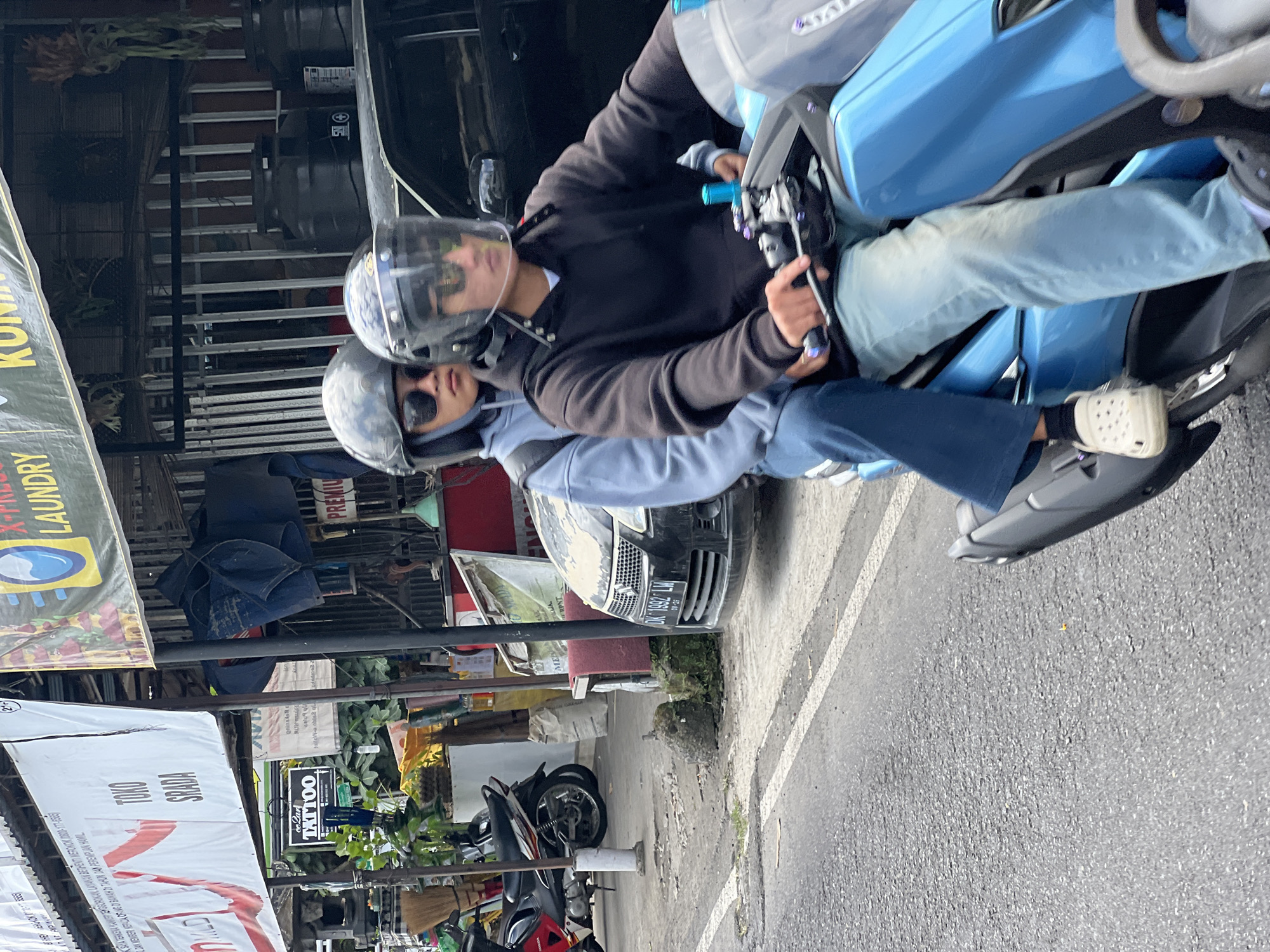 In Payangan, Indonesia, two individuals are riding a blue scooter on an asphalt road. The rider in front wears a black hoodie, light blue jeans, and a dark helmet with a clear visor. The passenger behind wears a light blue hoodie, dark blue pants, and a dark helmet. Both are wearing sunglasses. The scooter has a light gray body with blue accents. In the background, a black car with a license plate that reads "DK 1992 LW" is parked. Several businesses are visible, including one with a banner advertising "LAUNDRY" and another with a sign for "TATTOO." A red motorcycle is parked near the tattoo shop. The environment appears to be a bustling street with shops and parked vehicles.