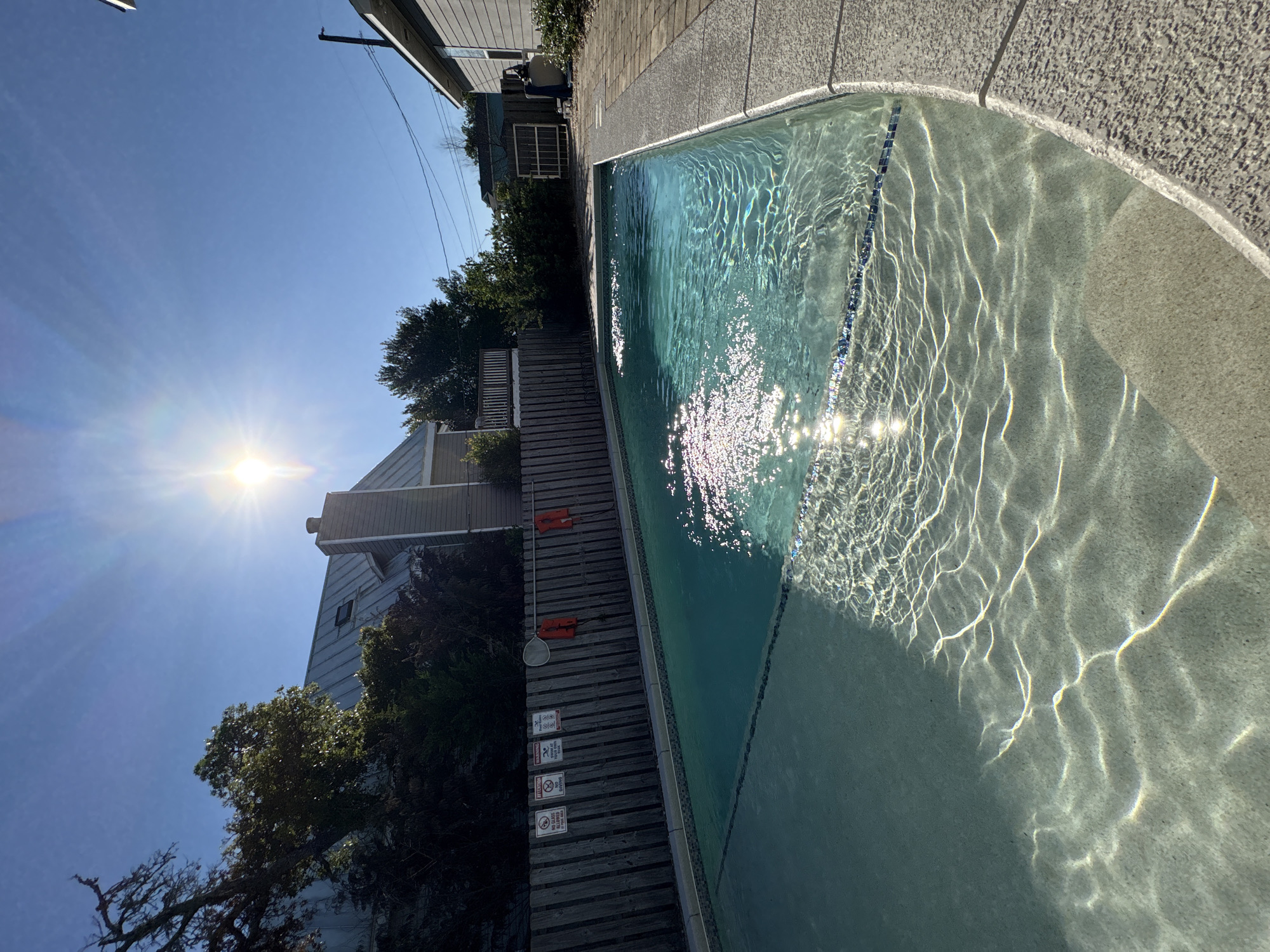 The image shows a partially visible swimming pool adjacent to a wooden deck and foliage. The pool water reflects sunlight, creating a shimmering effect. Safety equipment, including orange flotation devices and a pool skimmer, are visible on the deck. Posted signs with warnings and regulations are present on the deck. Houses and trees surround the pool area. The location appears to be Panama City Beach, United States of America.