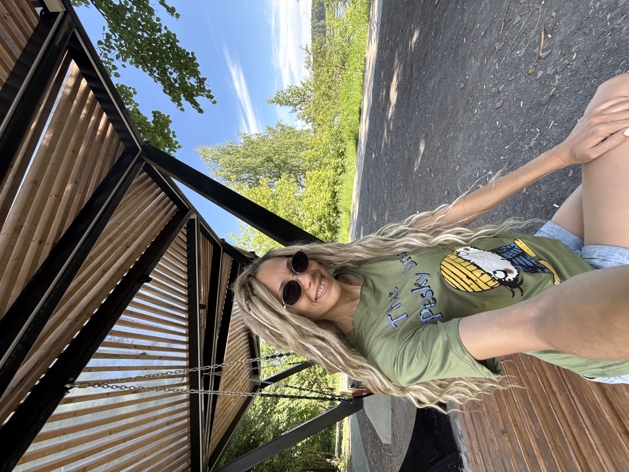 A woman with long, blonde dreadlocks and sunglasses smiles at the camera while sitting on a wooden bench under a covered structure. She is wearing a green t-shirt with a cartoon image and text. The background shows trees, a body of water, and a blue sky with clouds. The scene appears to be an outdoor recreational area with a swing set nearby.