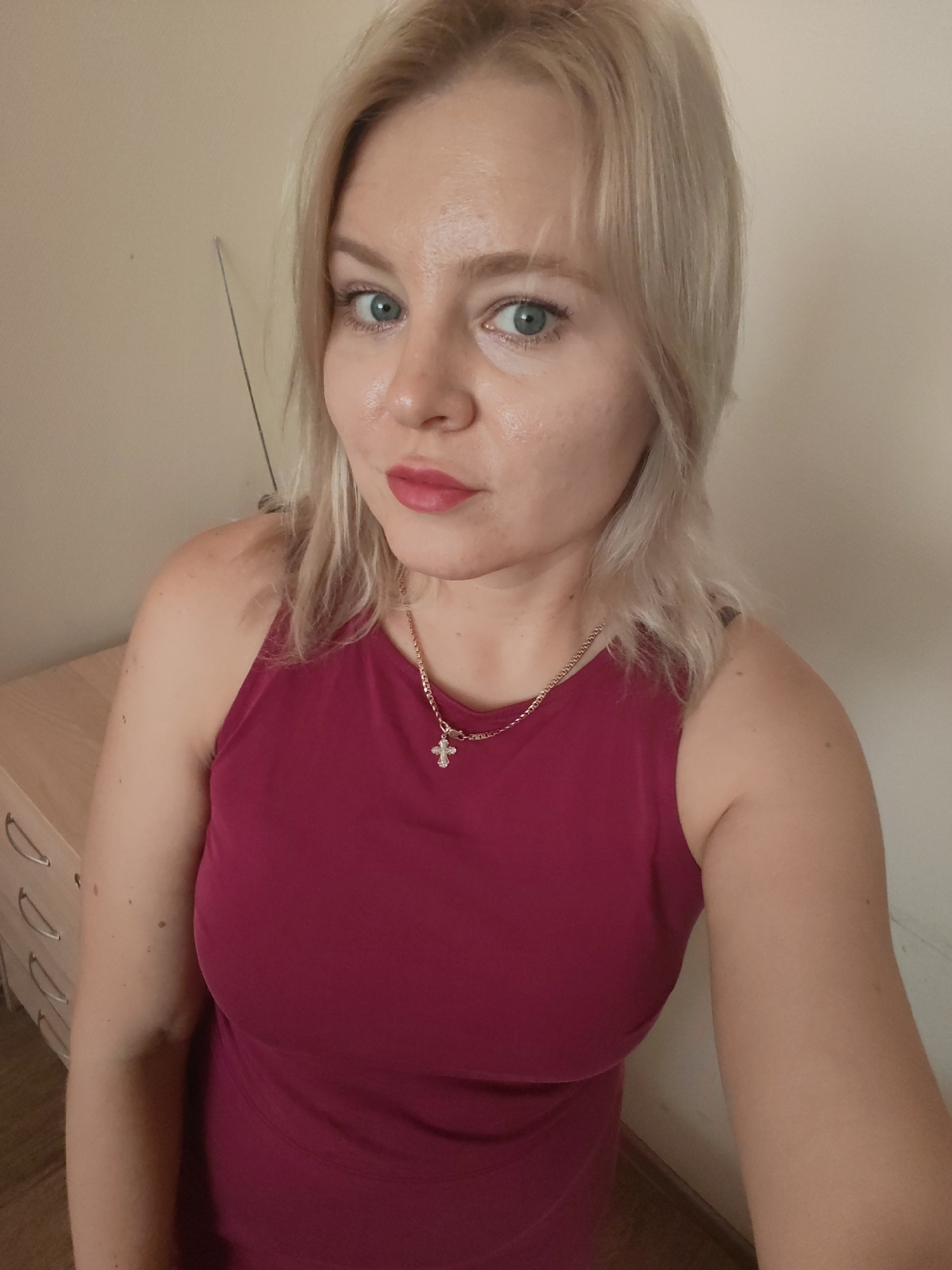 A young Caucasian woman with blonde hair and blue eyes is shown in a selfie. She is wearing a dark pink sleeveless top and a gold chain necklace with a cross pendant. Her makeup includes red lipstick. The background is a plain, light-colored wall. To the left of the woman, part of a wooden dresser with curved handles is visible. The overall setting appears to be indoors, possibly a home environment. The image was taken in the city of Izhevsk, Russia.