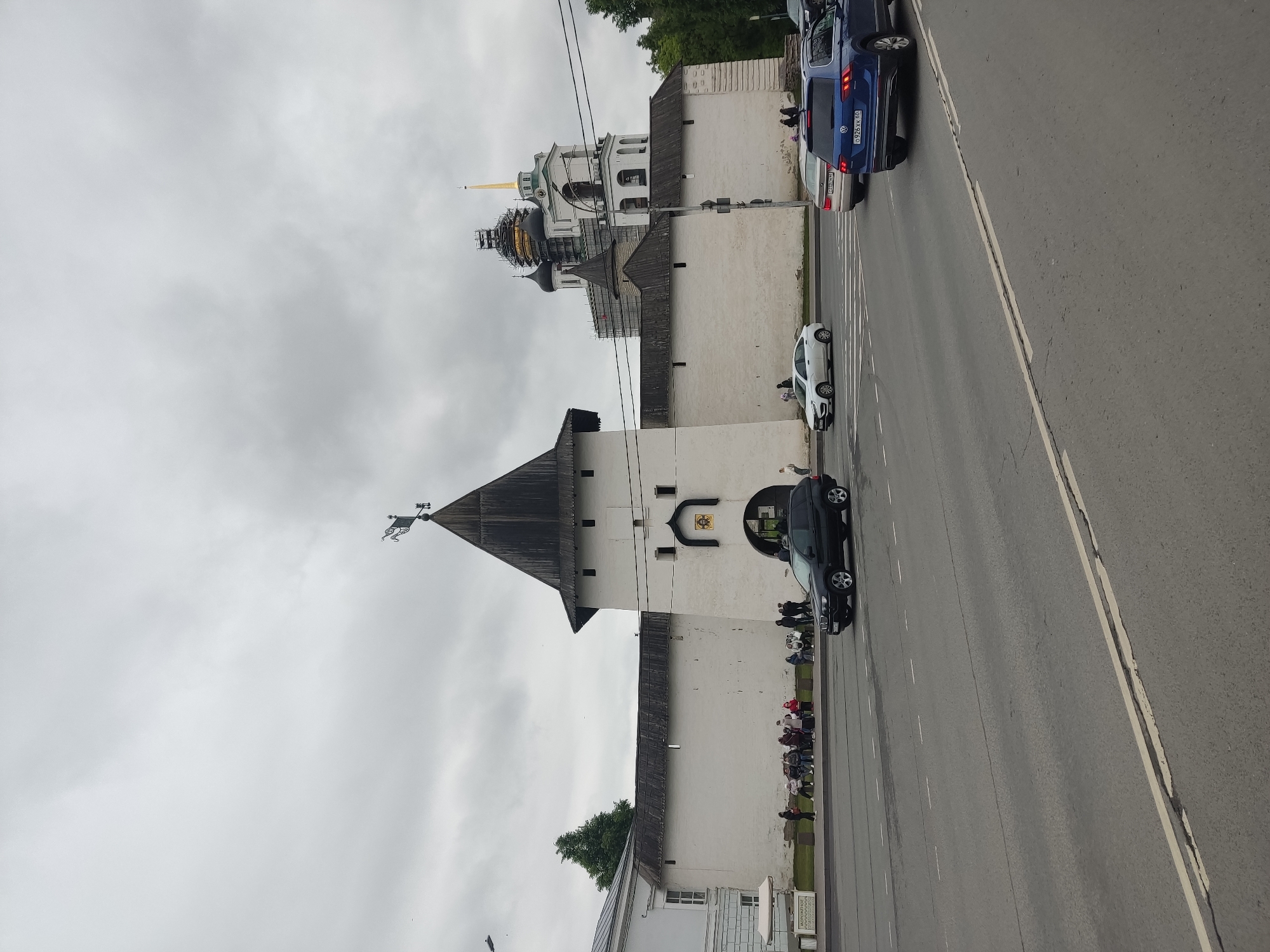 The image depicts a cityscape in Pskov, Russia, featuring a section of the city's ancient fortress walls. These white-stone walls are capped with dark wood and topped with towers, one of which is distinctive due to its pointed, dark roof and weather vane. A complex of buildings is visible behind the wall, including a multi-tiered structure with gold domes undergoing restoration. A group of people is gathered near the base of the wall, appearing to be tourists or sightseers. A road runs adjacent to the wall, with several cars parked or moving along it. The sky is overcast, casting a muted light on the scene.