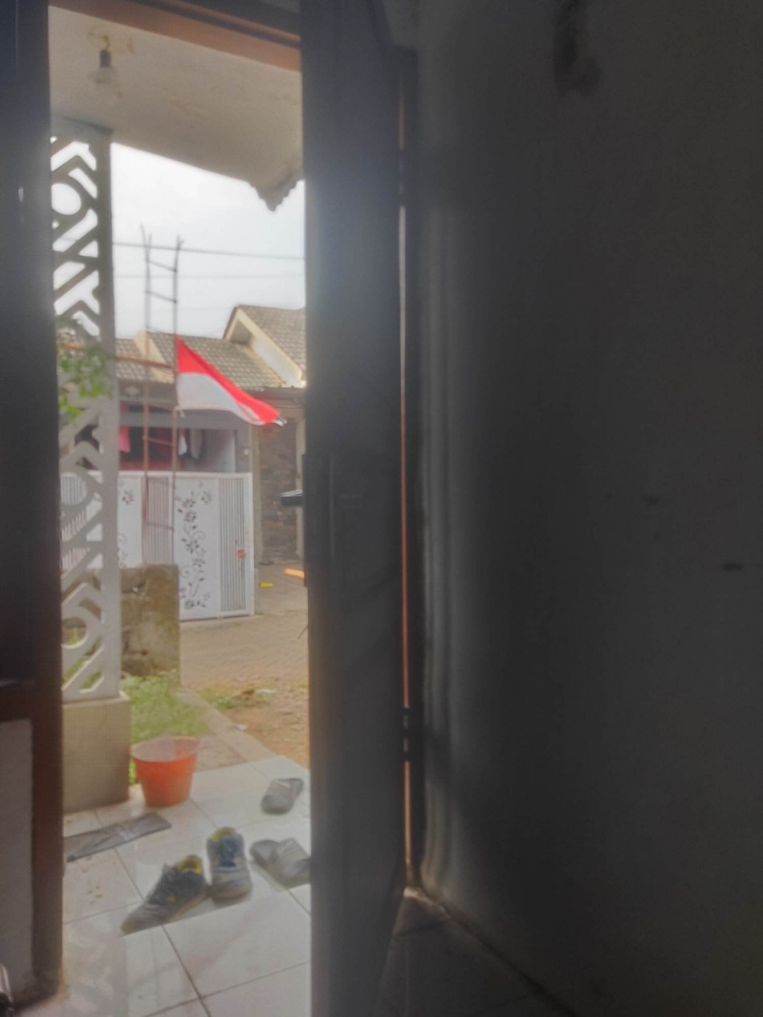 The image shows a view through an open doorway into a residential area in Antirogo, Indonesia. In the foreground, on a tiled floor, are a pair of sneakers and two pairs of sandals. An orange bucket sits near a decorative concrete pillar. In the background, a red and white Indonesian flag flies from a pole in front of a house with a tiled roof. A white metal gate with decorative floral patterns is visible. The overall scene suggests a domestic, outdoor setting on a slightly overcast day.