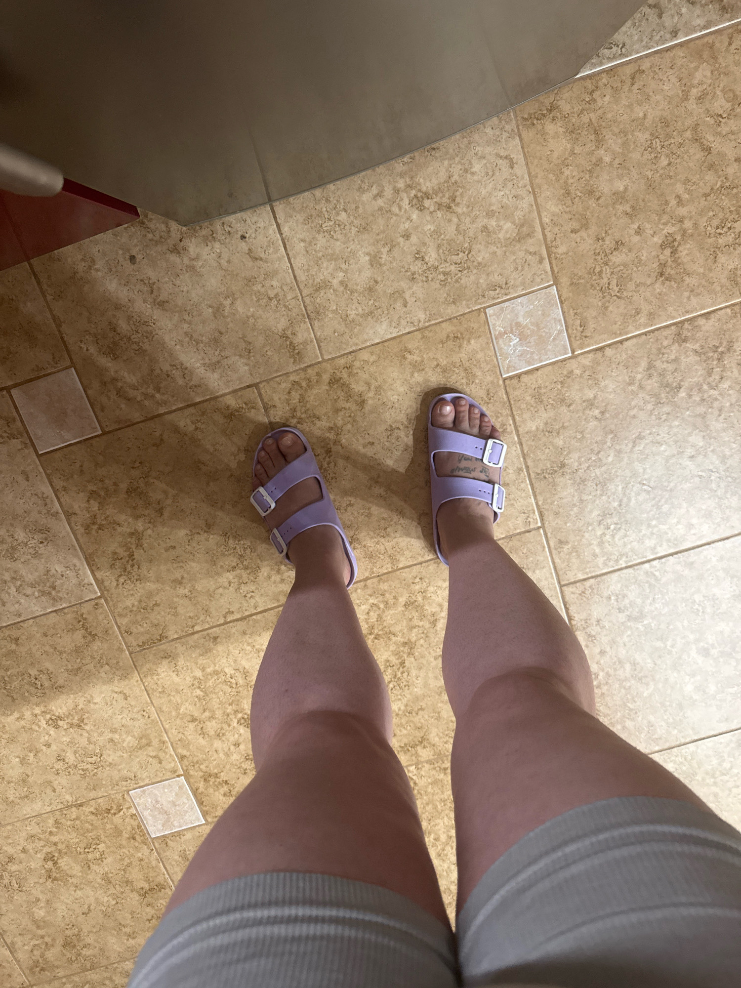 The image shows a pair of light purple Birkenstock sandals on a person's feet, standing on a light brown tiled floor. The person is wearing light grey shorts. The floor is made of square tiles with a mottled pattern, and there is a small, lighter colored square tile in the center of the frame. The scene appears to be indoors, possibly in a kitchen or bathroom, given the flooring. There is no visible indication of the specific location within the city of Hrodna, Belarus.