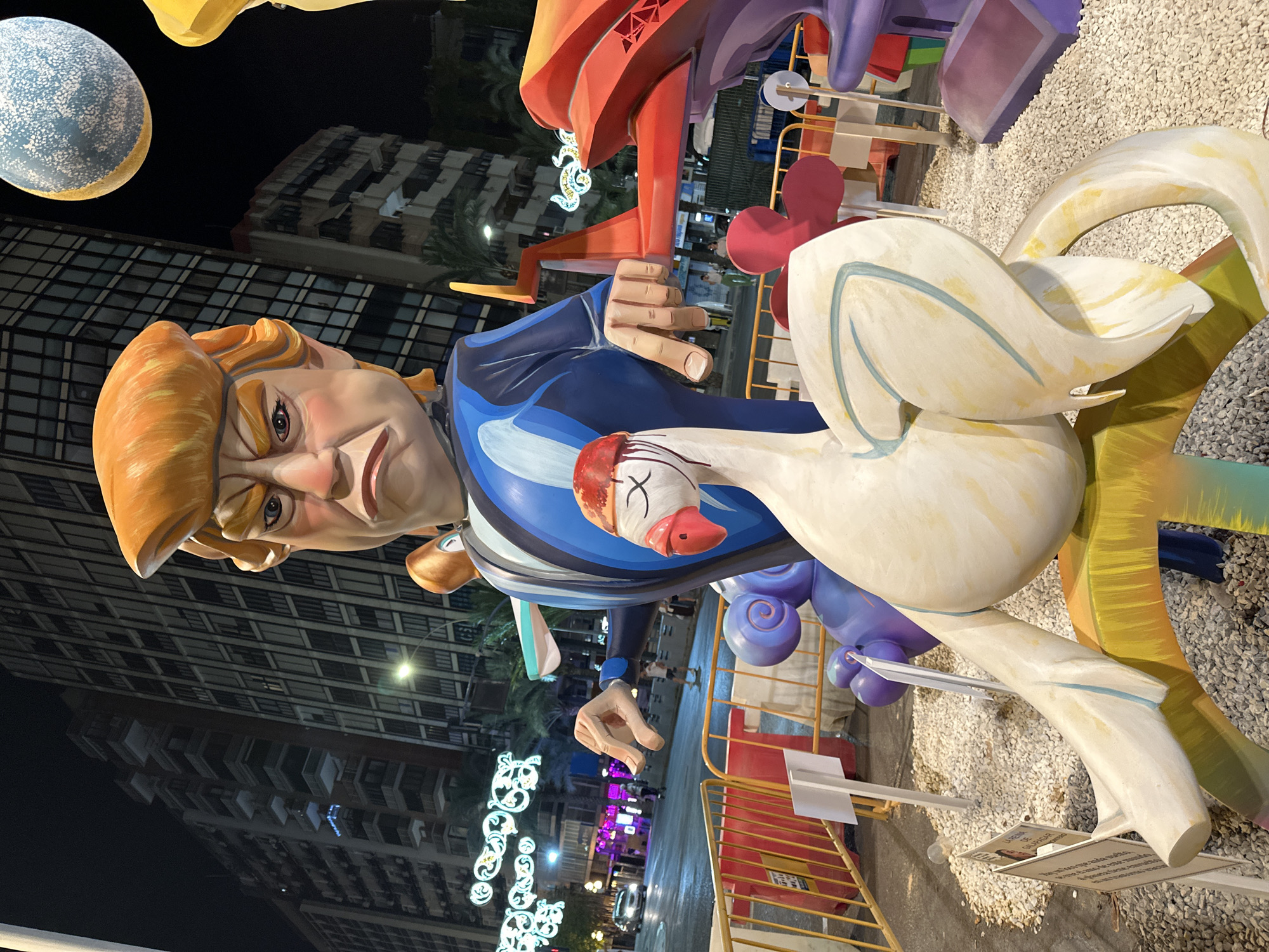 The image shows a nighttime outdoor scene in Alicante, Spain, featuring large, colorful figures. In the foreground, a white swan-like figure with a red hat is prominently displayed, resting on a multi-colored base. Behind it, a giant caricature of Donald Trump, with his characteristic orange hair and stern expression, is depicted wearing a blue suit. His hand is extended, pointing towards another large, abstract red sculpture. In the background, tall apartment buildings line a street, illuminated by streetlights and decorative lights. Several people are visible walking on the street, and cars are driving by. The figures are surrounded by gravel and temporary yellow and red barriers, suggesting a public event or exhibition.