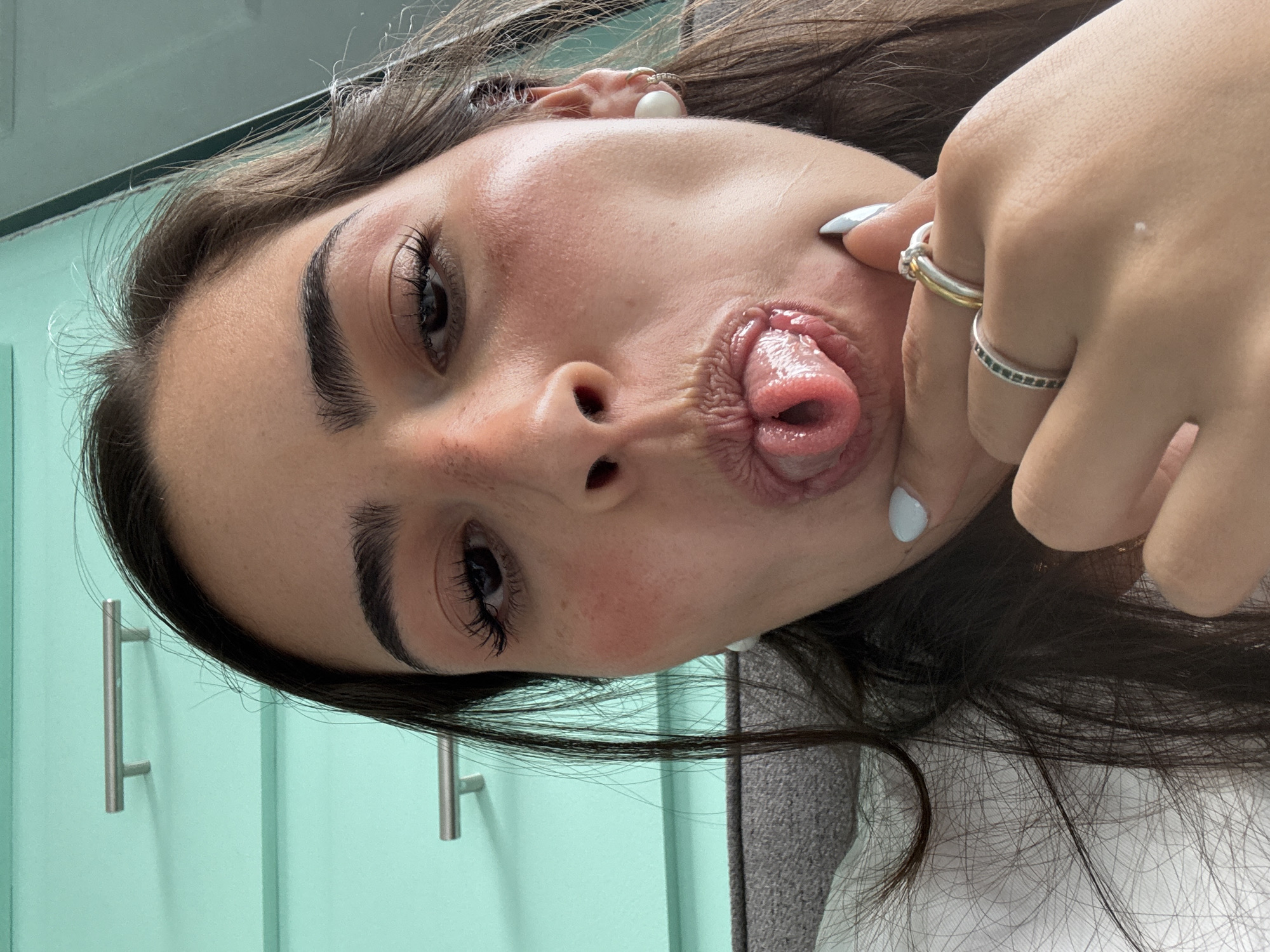 A woman with brown hair and olive skin is shown in a close-up selfie. She is making a playful face with her tongue sticking out and her cheek being pulled by her hand, on which she is wearing two rings. The background is blurred, but includes a light green wall with a silver handle, and part of a gray surface. The location is not evident from the image; therefore, it is not possible to determine if it is in San Bernardino Tlaxcalancingo, Mexico.