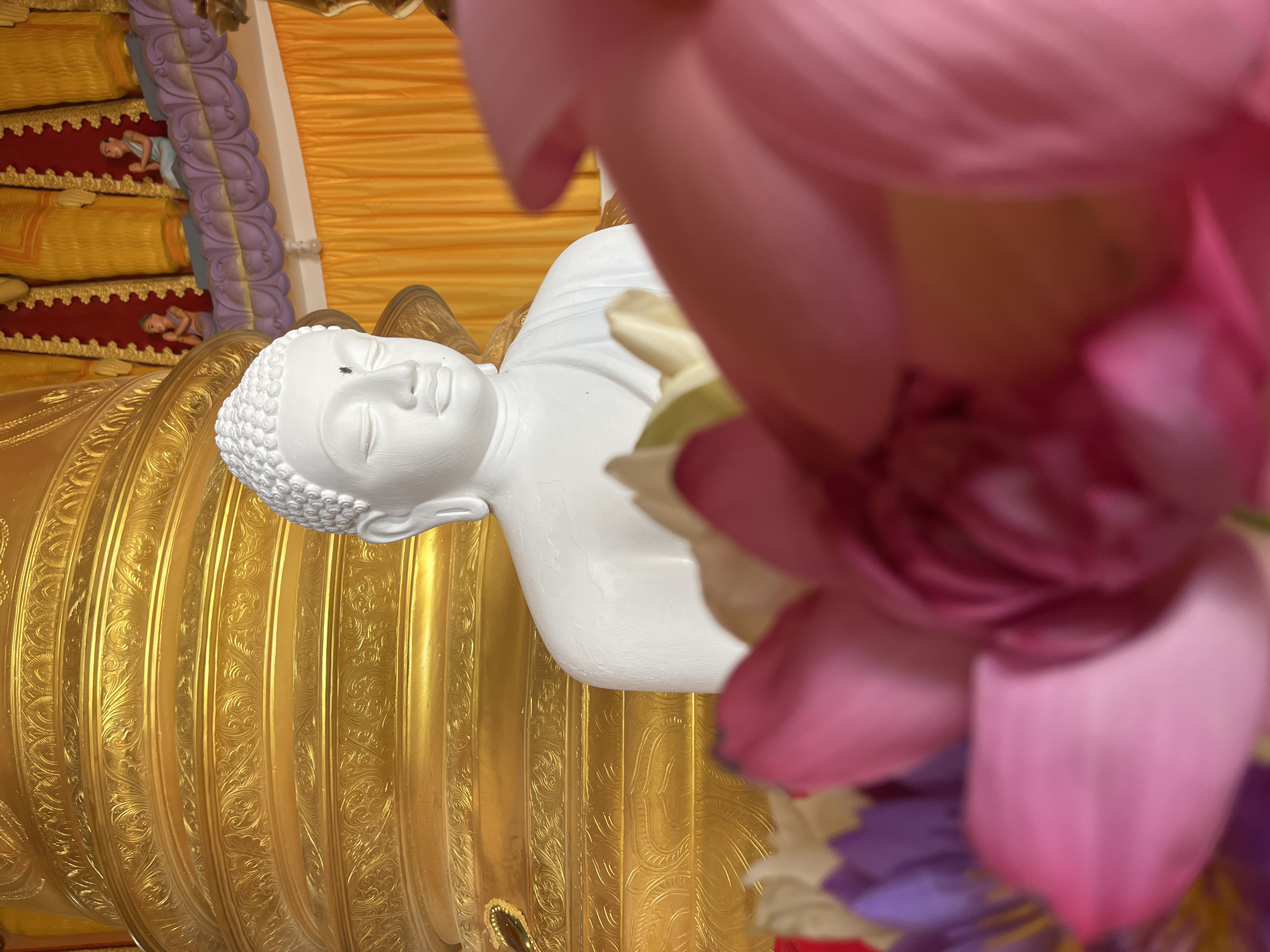 A close-up, eye-level shot shows a white statue of Buddha, its head slightly bowed, positioned to the right of a large, ornate golden pillar. The pillar features multiple horizontal bands with intricate, repeating relief patterns. Behind the Buddha statue and pillar, a yellow curtain hangs, and above it, several figures are visible, including two smaller statues in prayer. In the foreground, out of focus, are several lotus flowers in shades of pink and purple, partially obscuring the Buddha statue and pillar. The image appears to be taken inside a temple or shrine. The location is specified as Walgama, Sri Lanka.