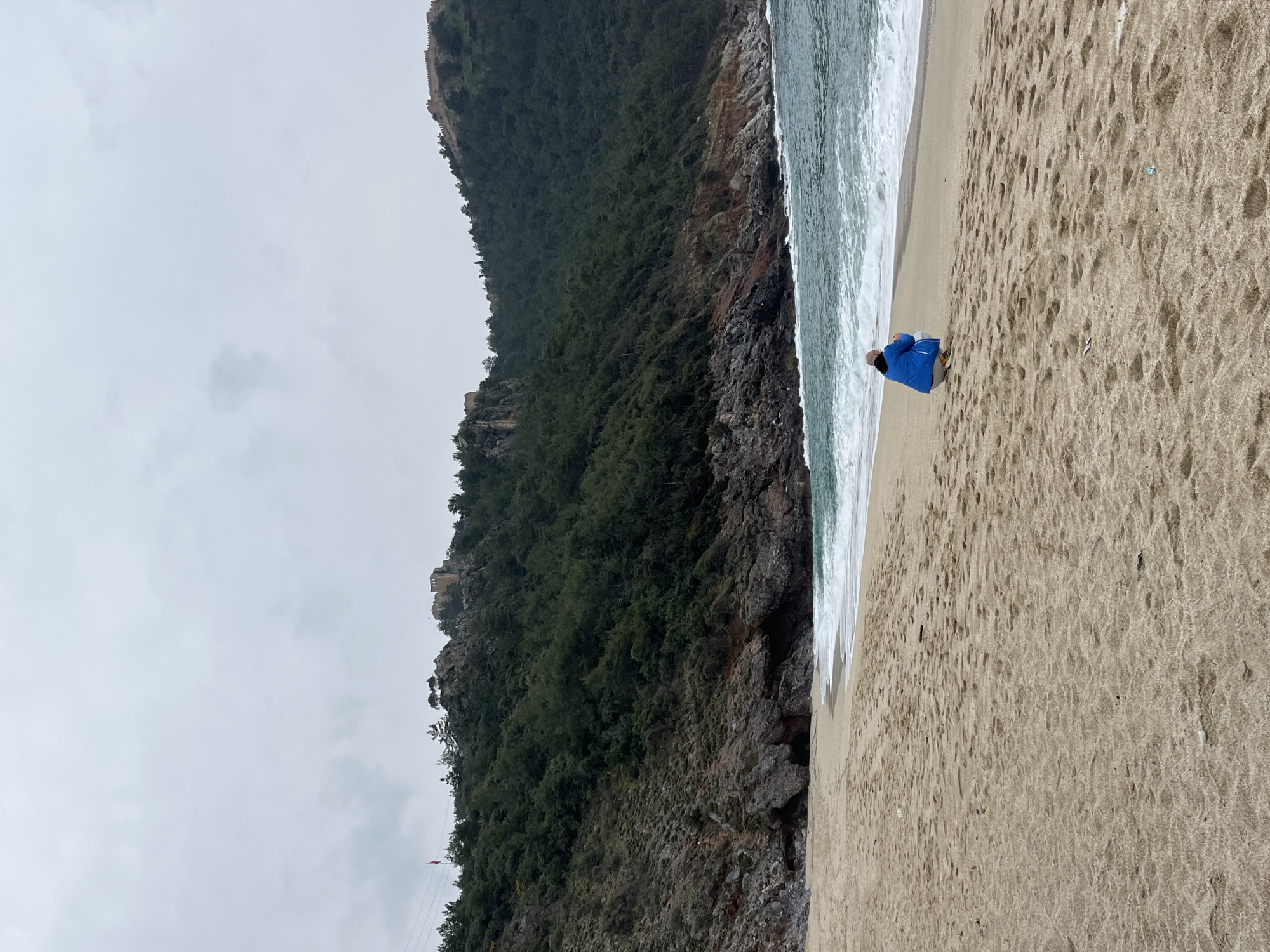 The image depicts a coastal scene in Alanya, Turkey, specifically a sandy beach situated along the base of the Alanya Peninsula, facing the Mediterranean Sea. The sky is overcast and grey.
In the foreground, a light brown sandy beach occupies the lower half of the frame. Numerous footprints are visible on the sand. A single individual, an adult male with a bald head, is positioned in a low squat or crouch near the water's edge, facing the sea. He is wearing a bright blue jacket, light-colored trousers, and light-colored shoes.
The midground features the sea, which exhibits a turquoise-green hue with white-capped waves actively breaking onto the shore. Beyond the beach, the coastline transitions into a rugged, rocky cliff face with various formations and some dark crevices at sea level.
In the background, a steep, densely forested hill rises prominently. This hill is covered primarily with dark green trees, suggesting a significant wooded area. Historical stone fortifications, consistent with sections of Alanya Castle, are visible on the upper reaches and along the crest of the hill. To the upper left, a red flag, likely the Turkish flag, is flying from a pole on the hillside. Power lines or similar cables are visible against the upper slope of the hill on the left side.