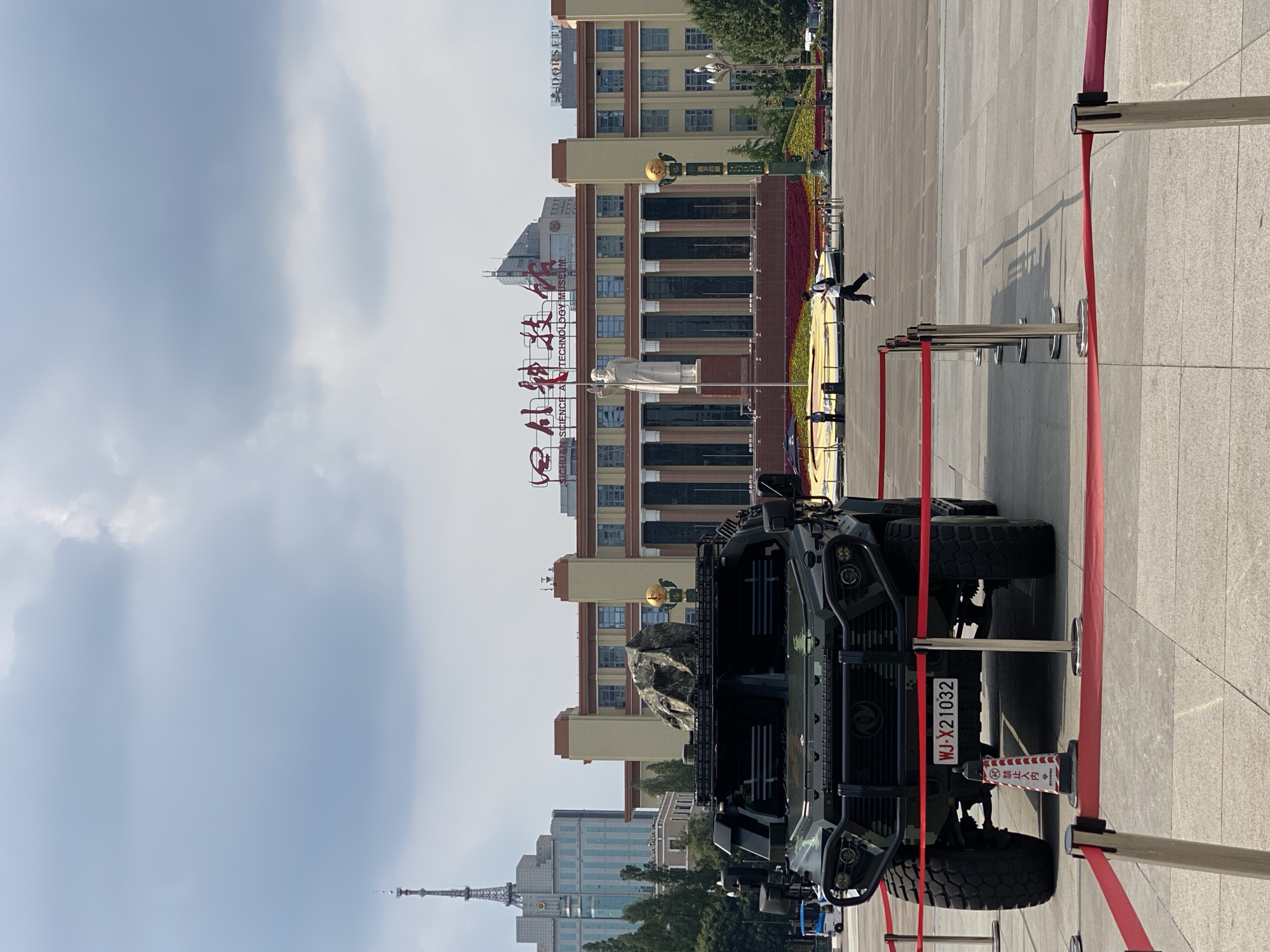 An outdoor medium shot captures a military-style vehicle positioned in a public square. The vehicle, dark green in color, is the primary subject, positioned in the foreground and taking up a significant portion of the frame. It is equipped with large, rugged tires and has a protective grille at the front. A license plate, reading "WJ-X21032", is visible on the vehicle. Behind the vehicle, a large building with a neoclassical architectural style stands, featuring a prominent statue of a man in front of it. The building is adorned with signage that appears to be in Chinese characters, with "SICHUAN SCIENCE AND TECHNOLOGY MUSEUM" legible in English. A person is walking across the square in the midground, to the right of the statue, wearing dark pants and a white shirt. In the background, various buildings and a tall radio tower are visible against a cloudy sky. Red velvet ropes and stanchions delineate a viewing area around the vehicle. The ground is paved with large square tiles. The overall environment suggests a public event or exhibition in a city square. The location is identified as Qingyang District, China.