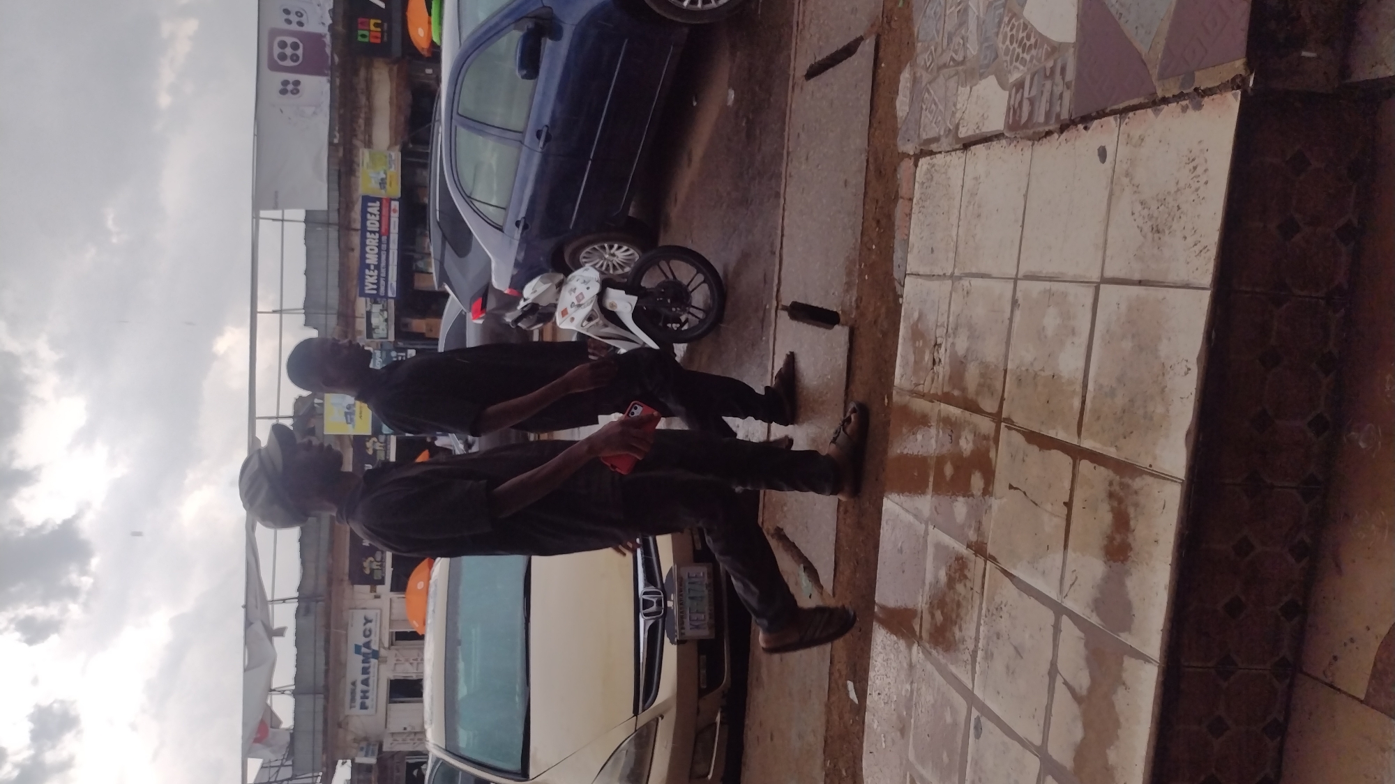 The scene depicts a street in Jos, Nigeria, with two men walking on a tiled sidewalk. The men are dressed in dark clothing. One man is holding a red phone. Parked cars line the street including a blue and gold sedan. A motorcycle is parked between the two cars. Behind the cars are commercial buildings featuring signs like 'Pharmacy' and advertising 'JYKE-MORE IDEAL CONCEPT ELECTRONICS'. The sky is overcast.