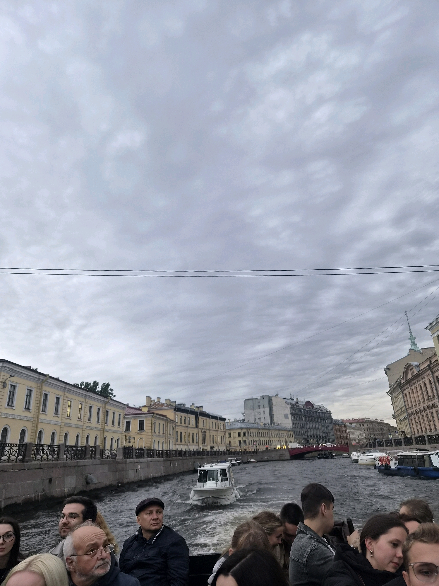 The image depicts a boat tour on a canal in Saint Petersburg, Russia. Several people are visible on the boat, some in the foreground looking forward and some further back. The boat is moving through the water, creating ripples. Along the canal, there are historic buildings with light yellow facades and arched windows. A white tour boat is seen in the distance, and other boats are moored along the canal. The sky is overcast with a textured cloud cover. Power lines are visible against the sky. The scene appears to be a daytime activity in an urban environment with historical architecture.