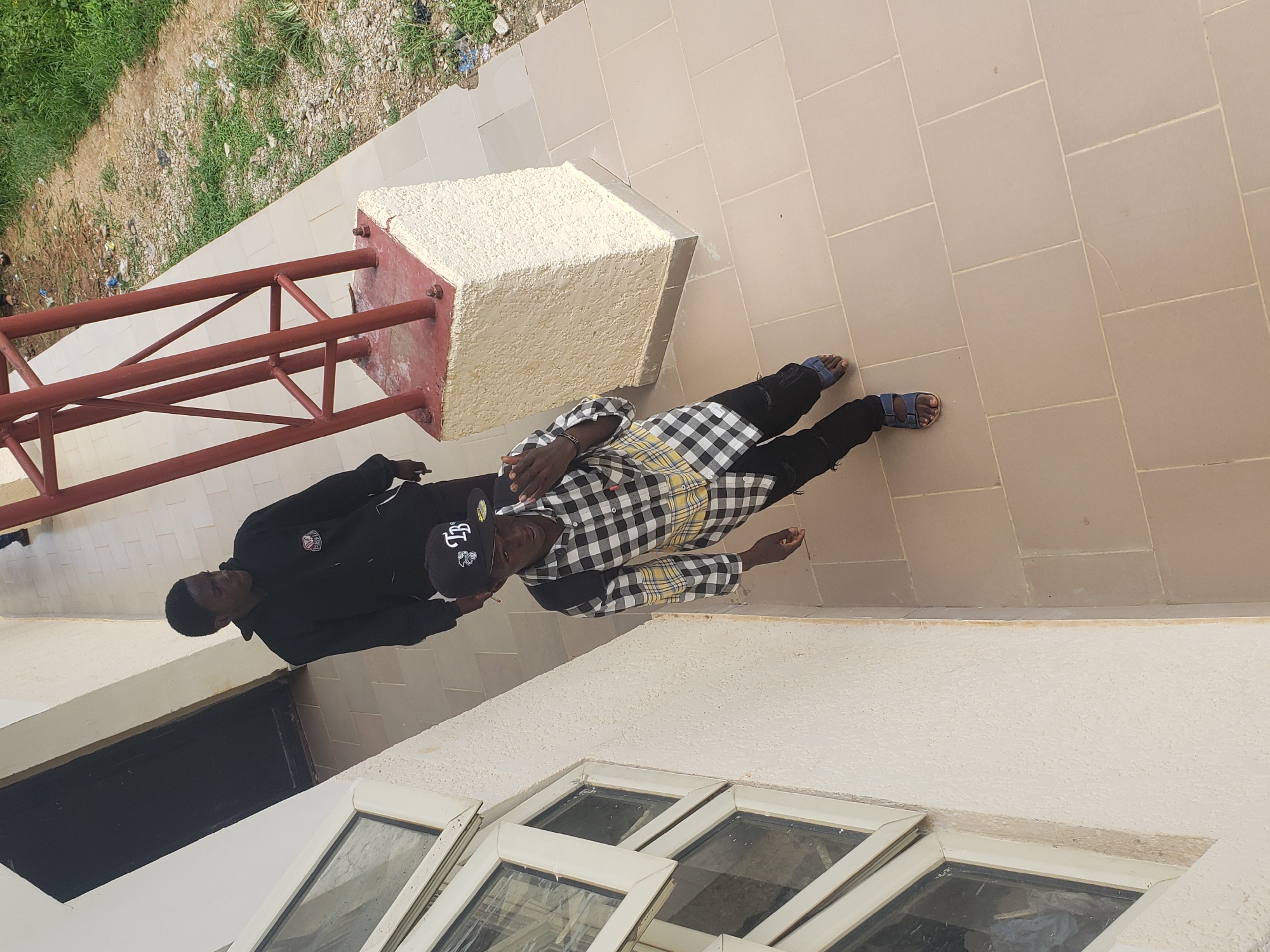 Two young men are standing on a tiled walkway outside a building. The man closer to the camera is wearing a black and white checkered shirt, black pants, and blue sandals. He is also wearing a black baseball cap with white lettering. He is holding a phone in his right hand and appears to be taking a selfie or recording a video. The second man, standing behind and to his left, is wearing a black hoodie and dark pants. He is looking to his right. To the left of the walkway is a cream-colored building with several large windows. To the right of the walkway is a patch of dirt and grass with some scattered debris. A red metal structure with a concrete base stands on the edge of the walkway. This scene is located in Gumel, Nigeria.