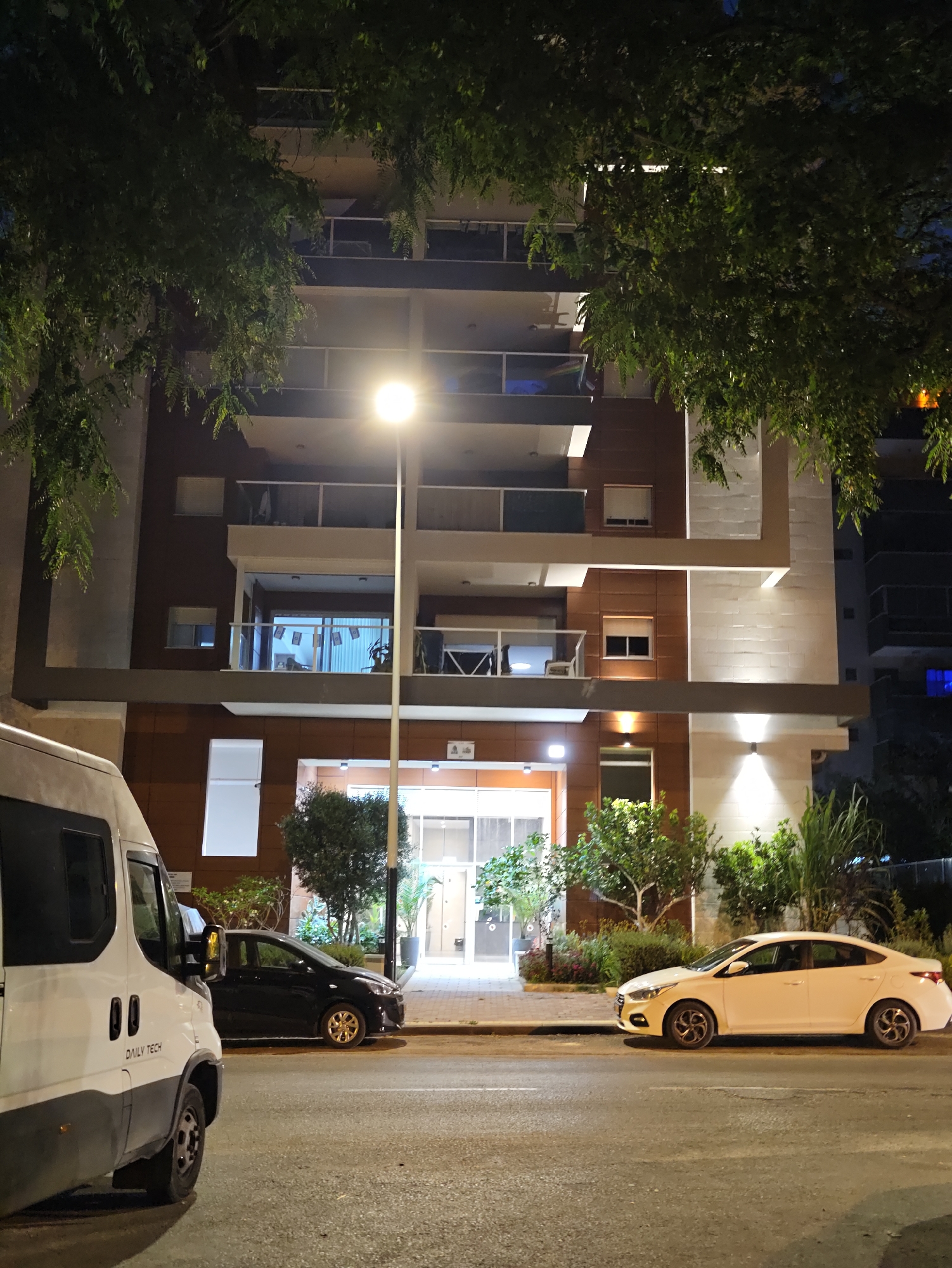 The image shows a building in Petah Tikva, Israel, at night. The building is modern with multiple stories, balconies, and large windows. There is a street lamp in front of the building, illuminating the scene. Several cars are parked in front of the building, including a white van on the left and a white sedan and a black sedan on the right. Trees and plants are visible around the building and on the balconies. The overall impression is a residential or commercial area at night.
