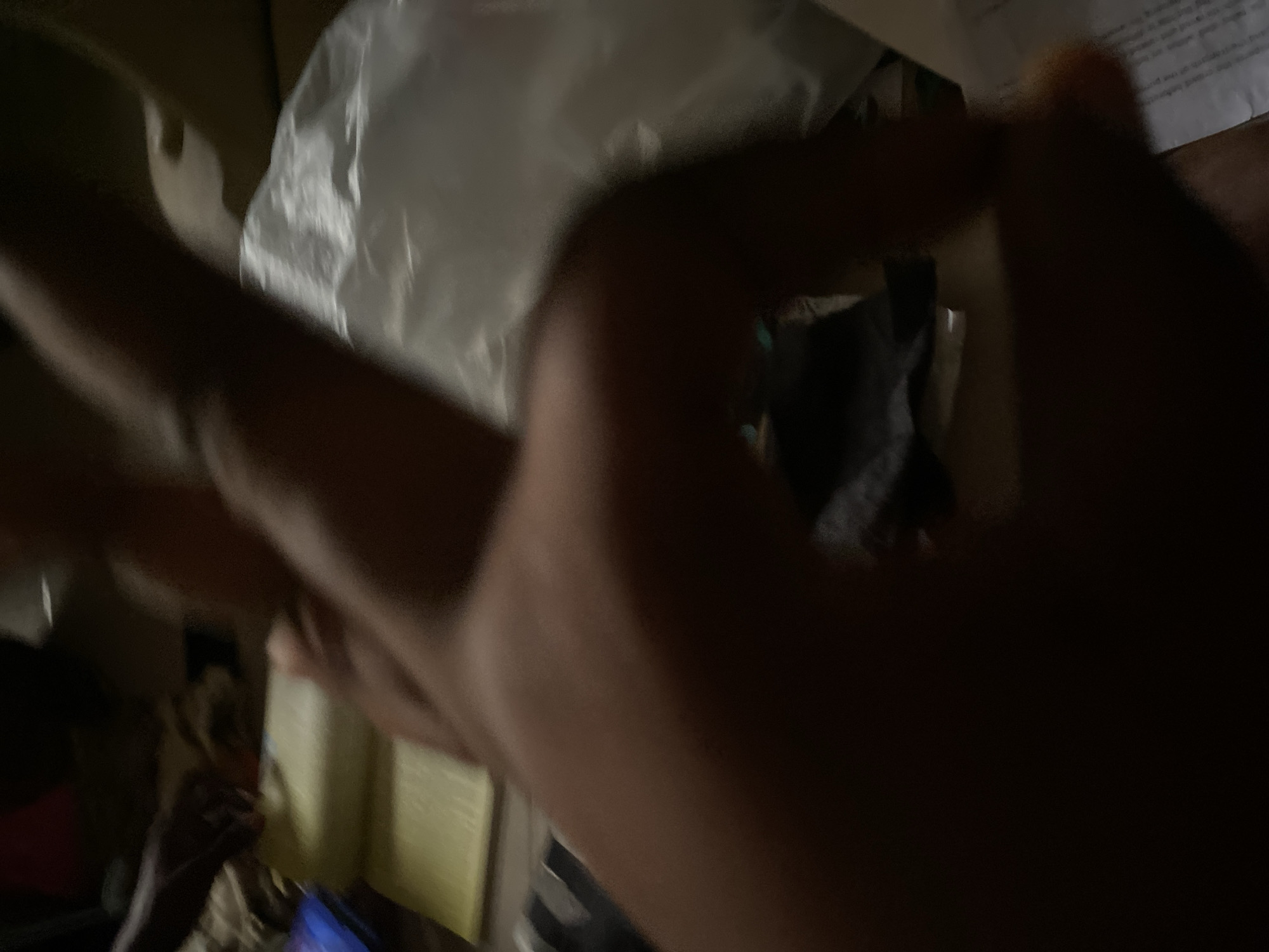 The image depicts a dimly lit indoor scene, likely a room, with a focus on a person's hand in the foreground. The hand appears to be positioned over a book or similar document with yellowed pages, suggesting it might be held open or turned. To the right of the hand, a crumpled white plastic bag is visible. The background is blurred and dark, with faint hints of other objects or furniture. The presence of a hand actively engaged with a book implies an activity like reading, studying, or note-taking. The location within Jauro Yinu, Nigeria is not discernible from the visual information provided.