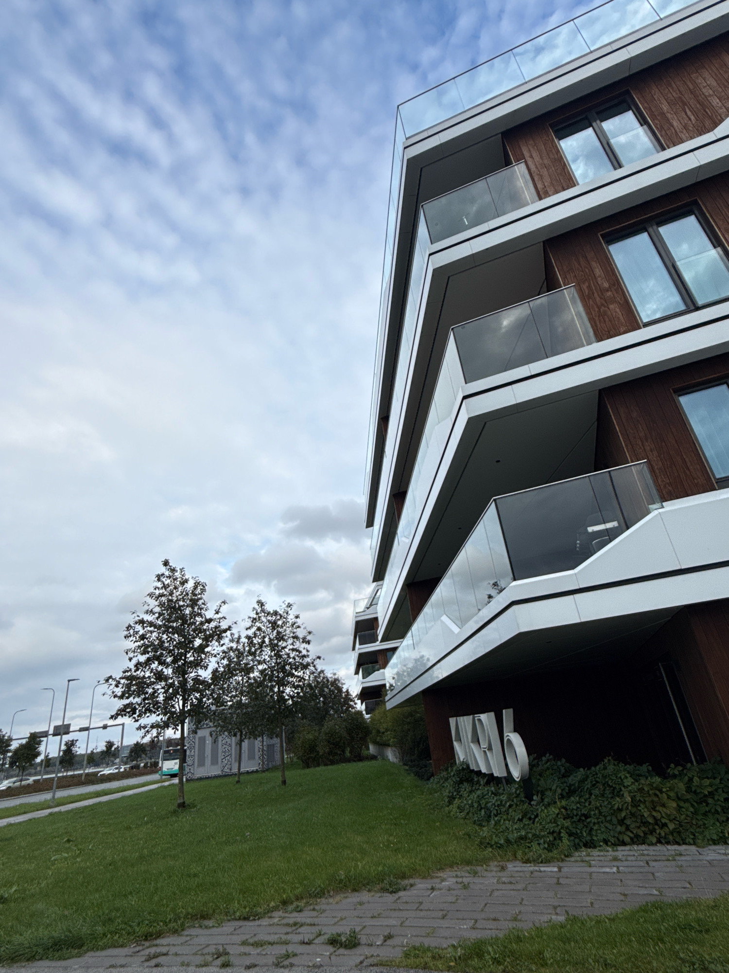 This is a low-angle, medium shot of a modern apartment building with a dark wood-paneled facade and white trim. The building has multiple balconies with glass railings and large windows reflecting the cloudy sky. To the left of the building, there is a grassy slope leading down to a sidewalk and a road. Several trees are visible on the slope, and in the distance, there are streetlights, a bus, and cars. The building has large white numerals "456" affixed to its lower section, and is surrounded by green shrubbery. The scene appears to be located in Tallinn, Estonia, as indicated by the building's signage.