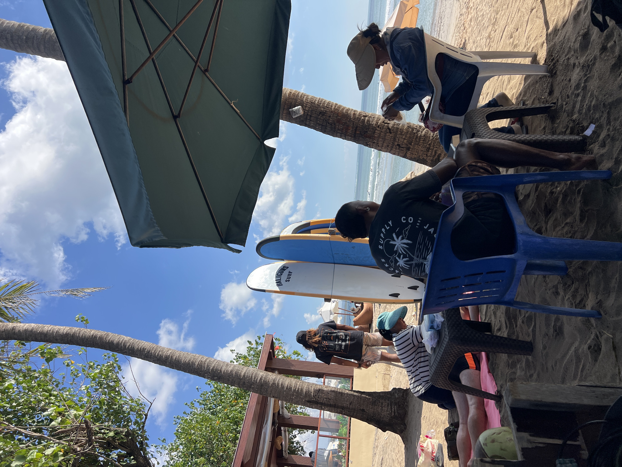 A group of people are relaxing on a sandy beach in Kuta, Indonesia, under palm trees and a large umbrella. Several surfboards are propped up behind them. In the foreground, a person in a striped shirt and blue hat is interacting with a small table. To their right, another person in a dark t-shirt sits in a blue chair. Further to the right, a person wearing a denim jacket and a wide-brimmed hat sits in a white chair, looking at their phone. The ocean and a few distant surfboards are visible in the background.