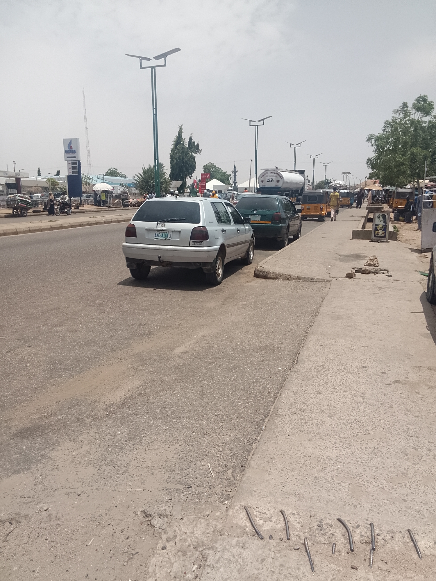 The image depicts a street scene in Pantame, Nigeria. A silver Volkswagen Golf and a dark green car are parked on the shoulder of an asphalt road. A white tanker truck is in the background, along with several yellow auto rickshaws (tuk-tuks) and pedestrians. A street lamp is visible in the background. The sidewalk is cracked and damaged, with exposed rebar. A sign is also visible on the sidewalk. In the far background, a fuel station can be observed. The sky is overcast.