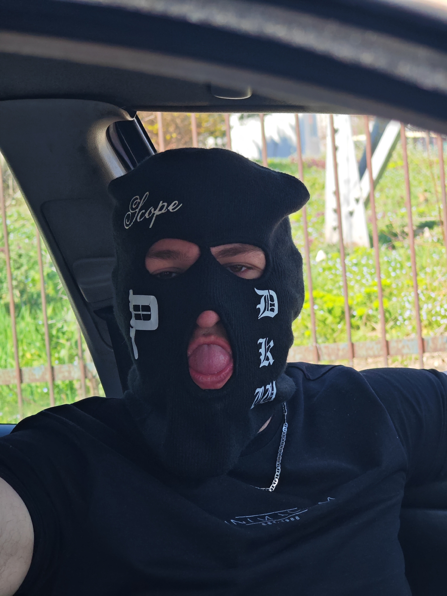 The image presents a head and upper torso shot of a single individual seated within a vehicle, likely in the front passenger seat. The vehicle's interior elements, including dark-colored headliner and pillar trim, are visible. The individual wears a black balaclava with cutouts for the eyes, nose, and mouth. The balaclava displays white text: "Scope" in script across the forehead, a stylized 'P' on the left cheek, and 'D K' stacked vertically on the right cheek. Visible through the balaclava are brown eyes and an open mouth with the tongue extended forward. The individual wears a black short-sleeved t-shirt, which partially reveals a white graphic including "EST. 1988" on the chest, and a silver-colored chain around the neck. Through the vehicle's window, an out-of-focus outdoor environment is visible, comprising green foliage and a rusty brown metal fence with vertical bars. The generic characteristics of the background do not provide specific landmarks for precise identification of a location within Shumen, Bulgaria. The scene is lit by natural daylight.