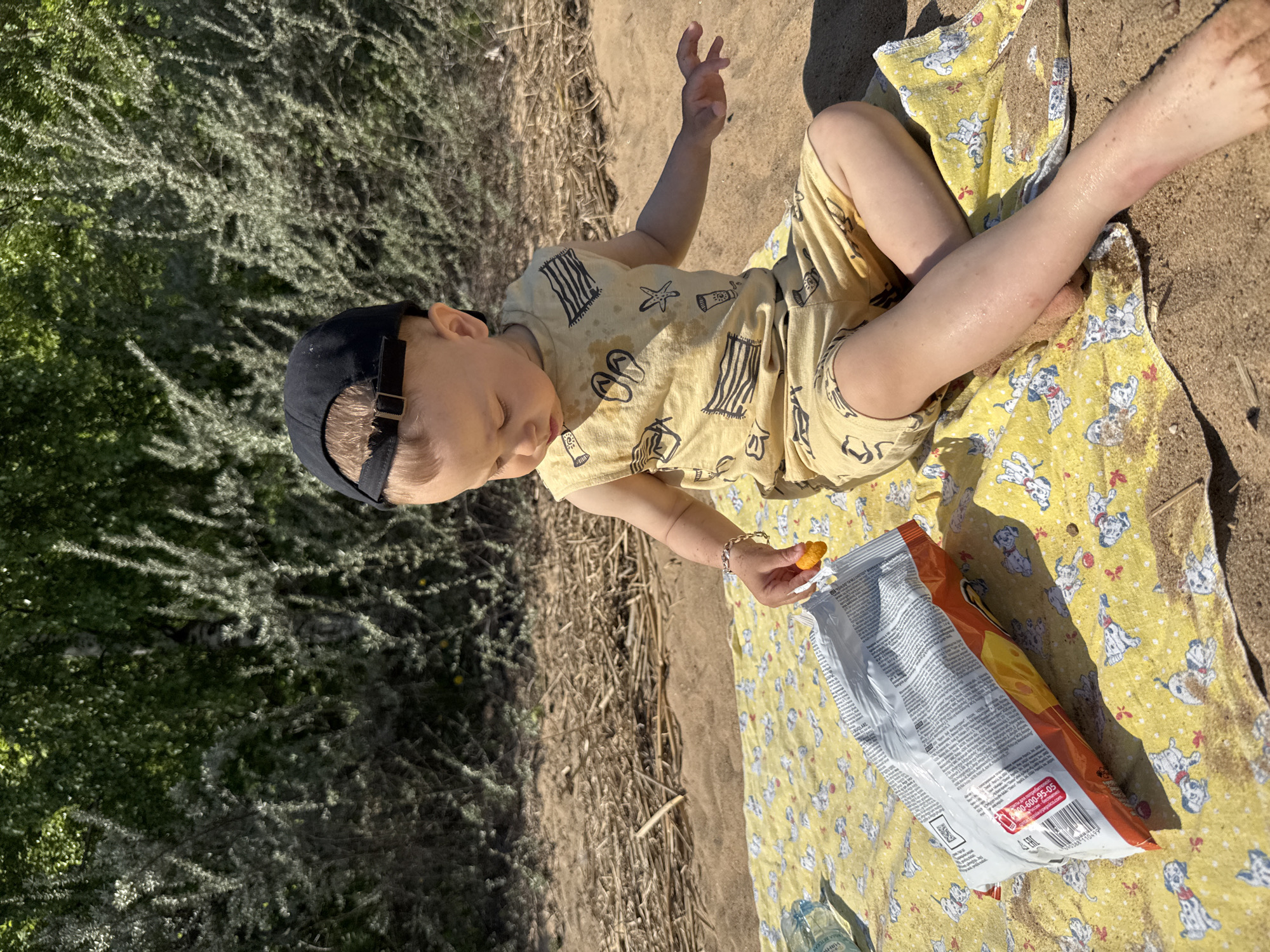 A young child is sitting on a yellow blanket patterned with dalmatians on a sandy surface, likely at a beach. The child is wearing a yellow outfit with beach-themed prints, a dark cap, and a bracelet on their wrist. They are holding a cheese puff. A bag of cheese puffs lies on the blanket near the child, and a bottle is visible near the bottom of the frame. The environment includes vegetation in the background.