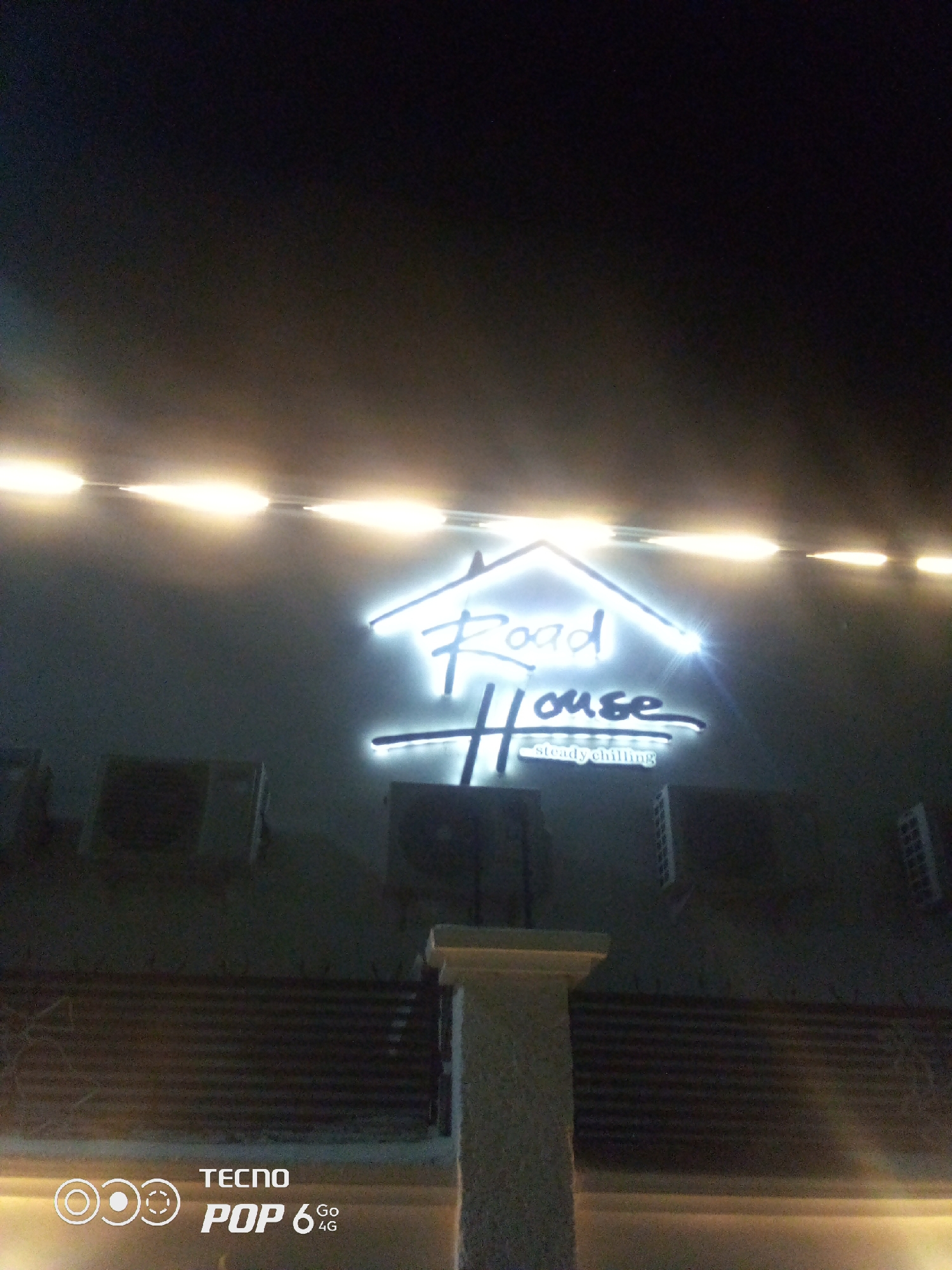 The image shows a lit sign that reads "Road House" in large, stylized font, with "steady chilling" written below in a smaller font. The sign is illuminated with white lights. Above the sign, a strip of yellow lights runs horizontally across the building facade. Below the sign, there are several air conditioning units mounted on the wall. In the lower part of the image, there is a column and a metal railing. The TECNO POP 6 Go 4G logo is visible in the bottom left corner. The scene appears to be an exterior shot at night, with the background dominated by darkness and the lights from the building. The specific location in Howayi, Nigeria, is not evident from the image itself.