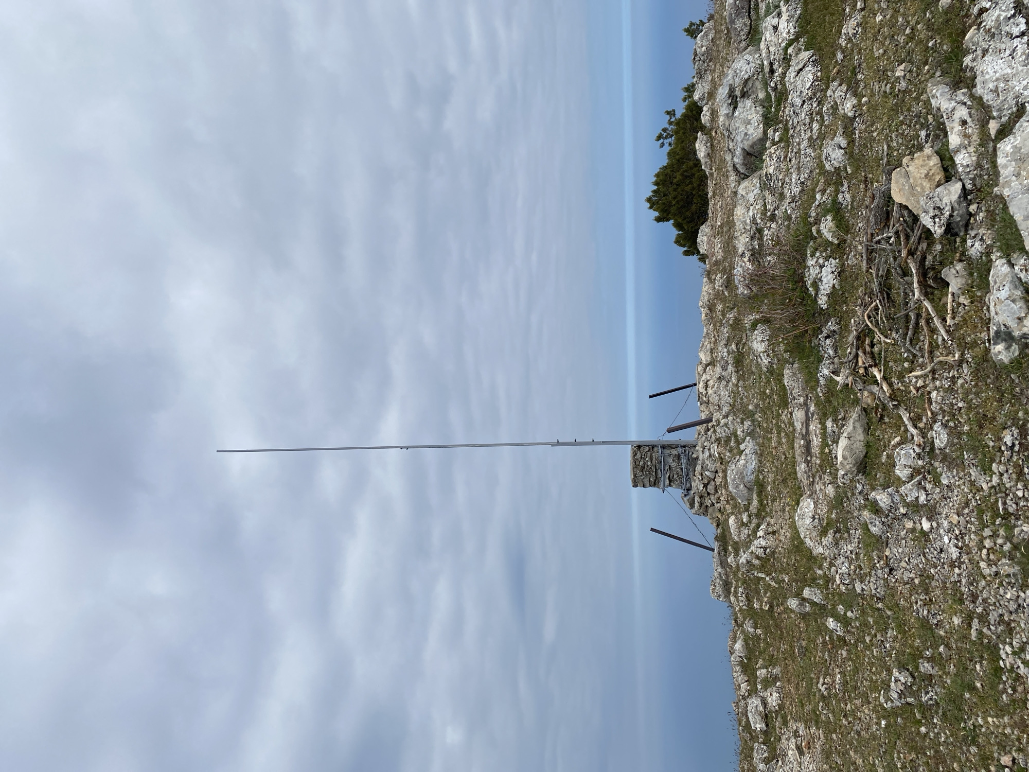 The image shows a rocky, mountainous landscape with a tall, slender metal pole extending vertically. The sky is overcast with a mix of light and dark clouds. The ground is covered with rocks, sparse vegetation, and some dry branches. A small stone structure is visible at the base of the pole. The horizon line is indistinct, appearing as a hazy blue band separating the sky from the ground. The location is Malyi Mayak, Ukraine.