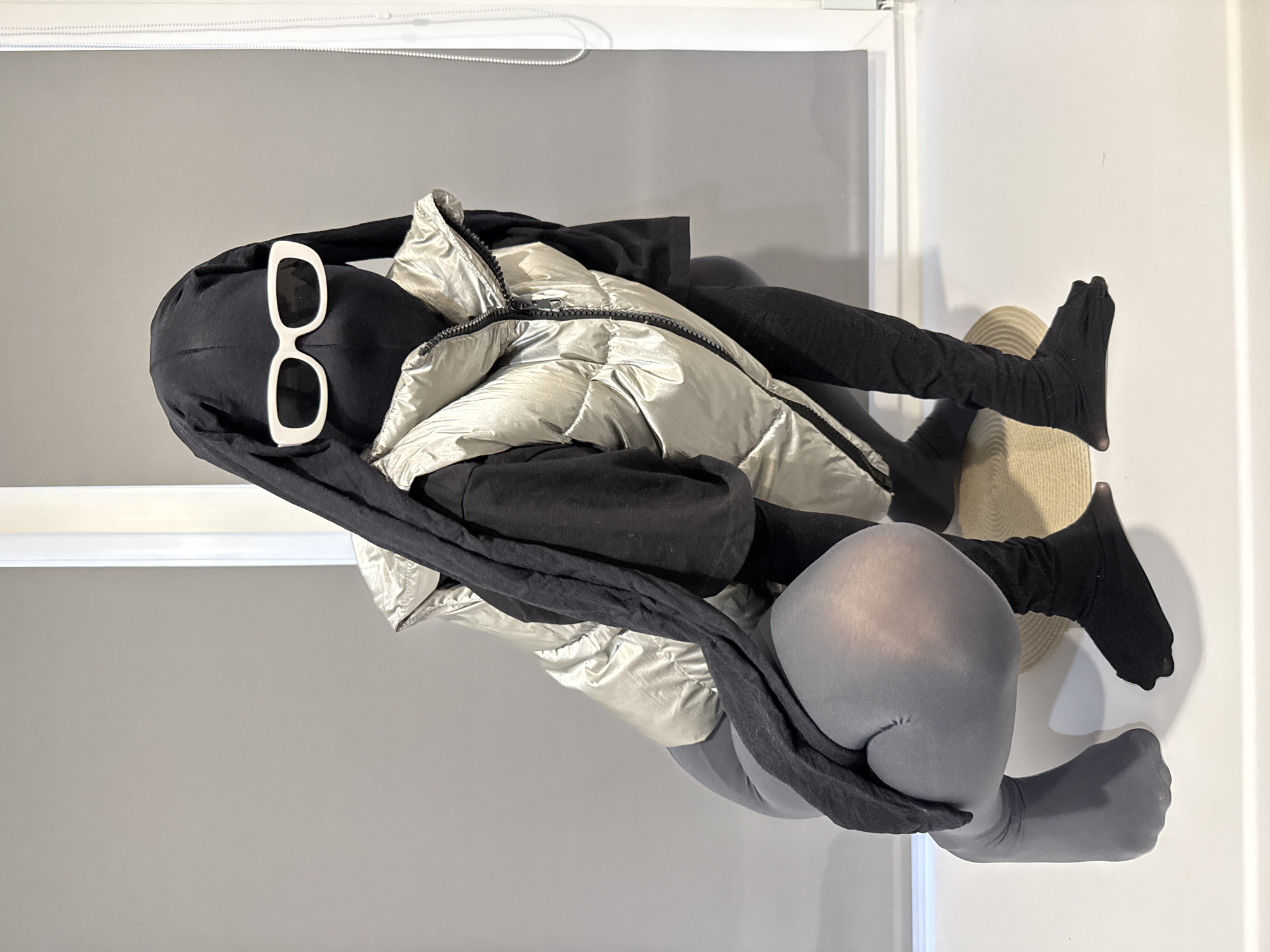 The image, taken in Rusanovo, Russia, features a faceless mannequin or display dummy posed in an indoor setting, showcasing a contemporary fashion ensemble. The mannequin wears a black full-body balaclava or bodysuit that covers its head and upper body. Prominently placed on its "face" are oversized white-framed sunglasses with dark lenses. Over the black base layer, a metallic, light silver or champagne-colored puffer vest with a visible zipper closure is worn, creating a layered and textured look.
A long, black piece of fabric, possibly a scarf or an integrated part of the bodysuit, is draped around the mannequin's neck and body. The lower half of the mannequin is clad in form-fitting, opaque grey tights or leggings. Its feet are covered in black, sock-like fabric, resembling soft boots. The mannequin is depicted in a seated or reclined position, with two light-colored, circular woven mats or hats positioned beneath its lower legs and feet, acting as props or a base.
The background consists of a plain wall, primarily light grey on the left and white on the right, suggesting an interior corner or a two-toned wall. A simple white horizontal trim or molding is visible along the top edge of the wall. The lighting appears bright and even, characteristic of an indoor environment. There is no visible text in the image. The overall aesthetic is modern, monochromatic, and emphasizes material textures and distinct shapes.