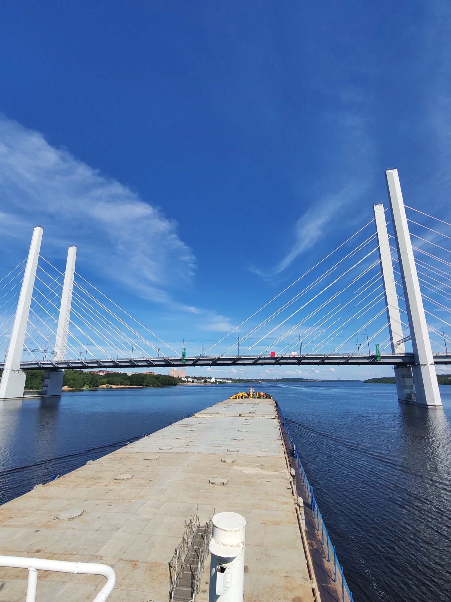 The image depicts a wide, bright blue sky with wispy white clouds. In the foreground, the deck of a barge is visible, leading towards a large, modern cable-stayed bridge. The barge's deck is covered in metal panels with circular hatches. A white railing is visible in the lower left corner. The barge appears to be moving forward, as indicated by the wake in the water.
The bridge is the prominent feature of the mid-ground, with its two distinctive white towers and numerous white cables extending to the bridge deck. The bridge spans a wide, calm body of water, likely a river or canal, with lush green trees and buildings lining the far bank. The sun is high in the sky, suggesting it is daytime, and the clear sky indicates fair weather.
In the distance, along the far bank, there are several buildings, including what appears to be a residential area and some industrial structures. A few small boats are visible on the water in the distance. The overall scene conveys a sense of journey and industrial progress. No visible text is present in the image. The location context provided is Cherepovets, Russia.