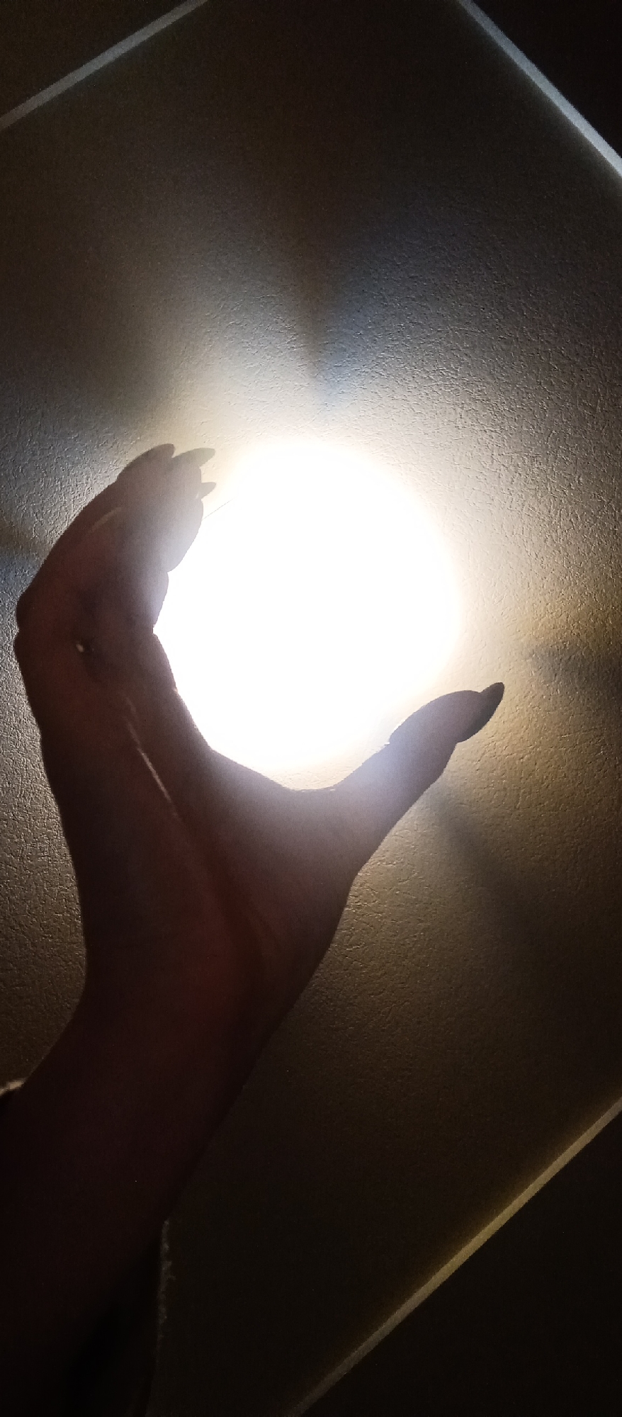 A hand is positioned in front of a light source on the ceiling, creating a silhouette effect. The background reveals a textured surface, likely a ceiling. The city location is unknown.