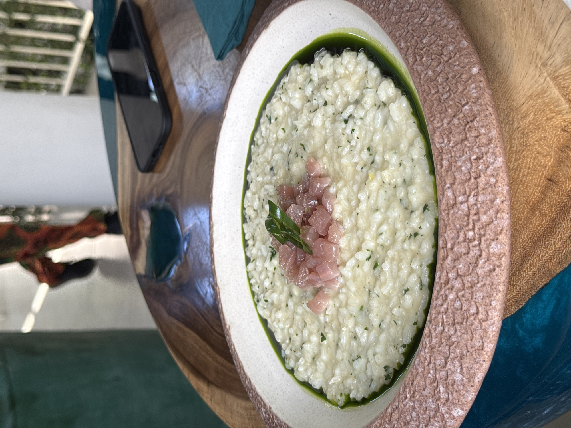 The image shows a plate of risotto, topped with diced fish and herbs, resting on a wooden table. A dark-colored smartphone sits next to the plate. In the background, a teal-colored material is visible. The location is North Kuta, Indonesia.