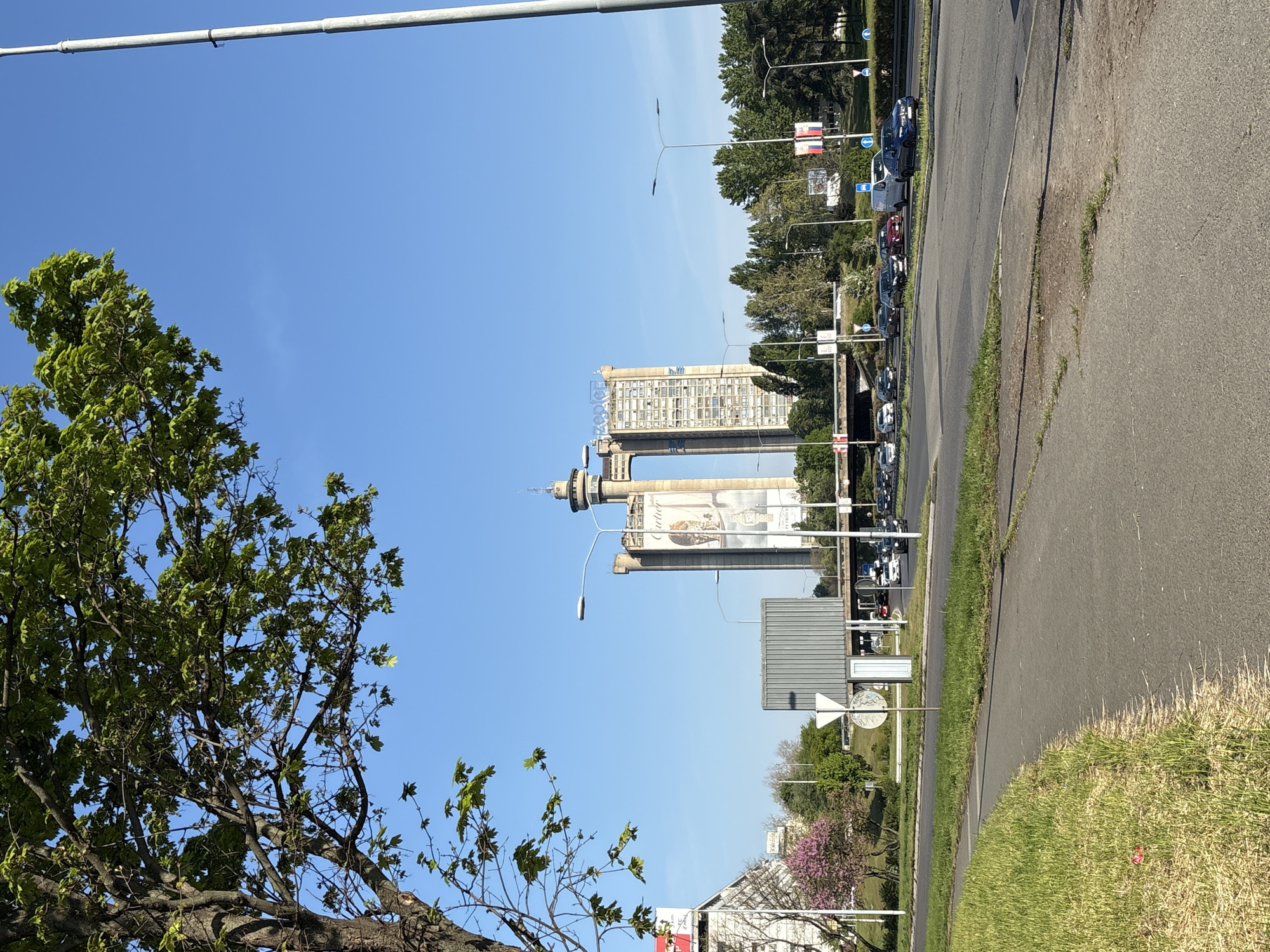 The image shows the Genex Tower in Belgrade, Serbia, taken from street level. The Genex Tower is a tall, brutalist-style building consisting of two connected towers, one being higher than the other, and connected with a bridge. One tower is residential, and the other is office space. It’s a clear day with blue skies. A leafy green tree is visible on the left of the frame, partially obscuring the view. A road runs along the right side of the frame, with parked cars along the far side. There are street lights along the roadside and various signs. A building in the foreground shows a store. The image is taken during the day.