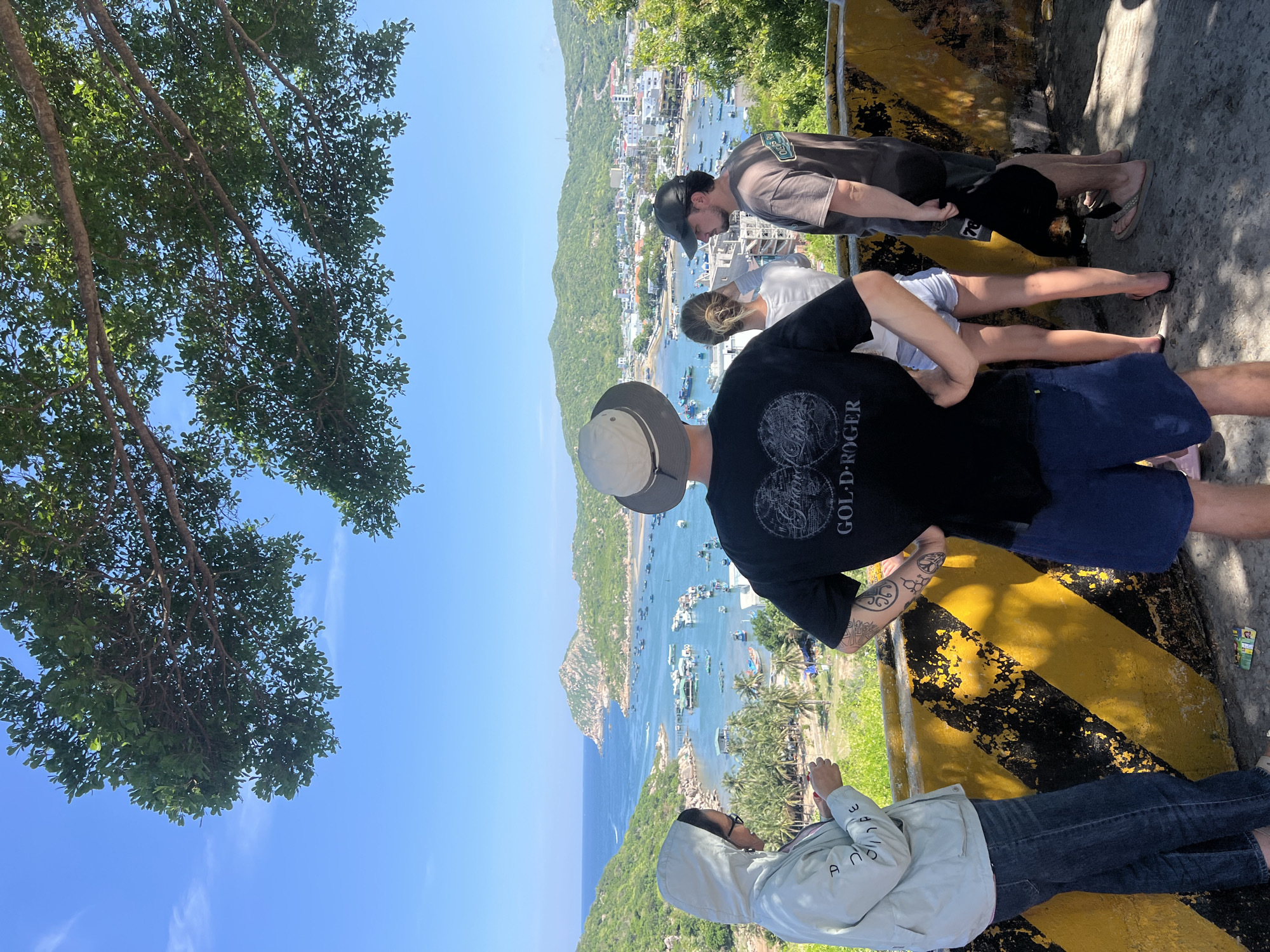 The image shows a group of four individuals standing on an elevated viewpoint overlooking a bay in Vĩnh Hải, Vietnam. The viewpoint has a yellow and black striped barrier. The individuals are positioned from left to right: a person wearing a light-colored hooded jacket and jeans, looking down at something in their hands; a person in the center wearing a black t-shirt with a graphic design and blue shorts, with their hands on their hips; a person behind them wearing a white t-shirt and denim shorts, facing away from the camera; and a person on the far right wearing a grey t-shirt and dark shorts, also facing away from the camera.
The bay below is filled with numerous small boats. In the middle ground, there is a village with buildings and more boats. The background features lush green hills and a clear blue sky with some light clouds. The foreground is dominated by the viewpoint and the people. The overall scene suggests a scenic overlook in a coastal Vietnamese town.
