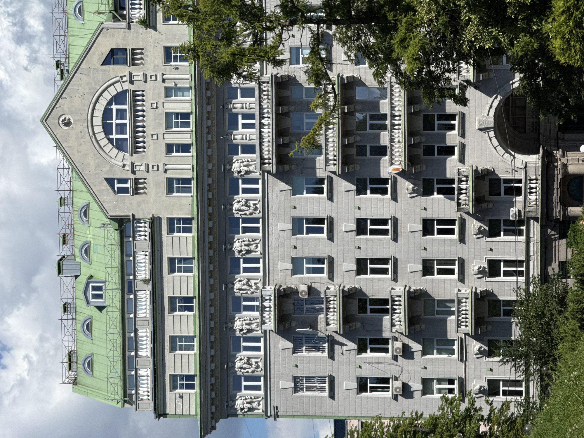 The image shows the facade of a historical building in Kyiv, Ukraine, characterized by its ornate gray stone architecture and a distinctive green mansard roof. The building features multiple stories with numerous windows, many of which are rectangular and symmetrically arranged. Several balconies, adorned with decorative balustrades, project from the facade. Notably, the upper floors display sculptures of human figures and decorative reliefs above the windows. The roofline is punctuated by dormer windows and a prominent arched window on the gable. Green trees partially obscure the lower portion of the building, suggesting an urban setting with a park or tree-lined street. The sky above is partially cloudy.
