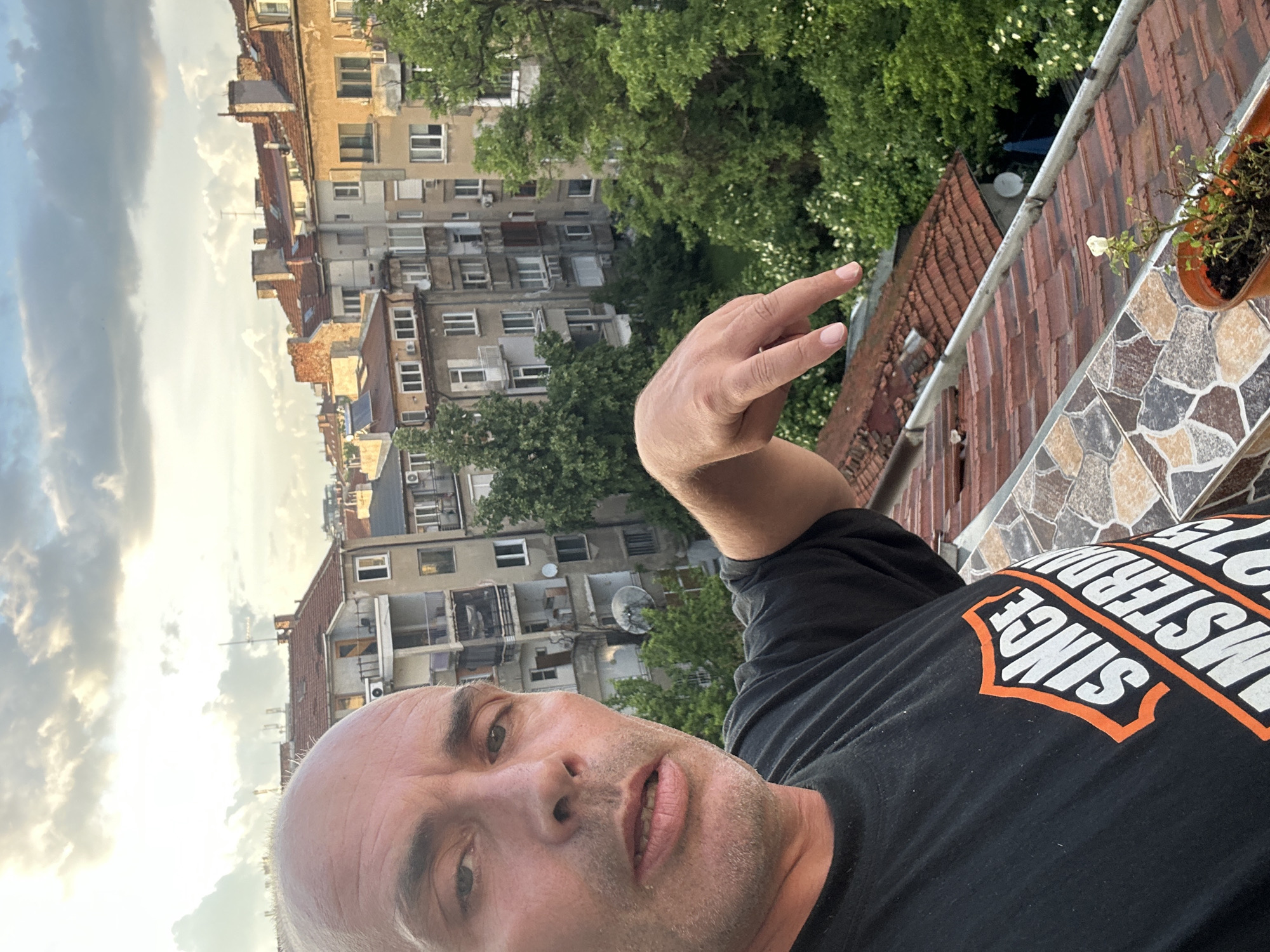 A man is taking a selfie in Sofia, Bulgaria. He is bald, wearing a black t-shirt with "SINCE 1973 AMSTERDAM" printed on it. He is outdoors on a rooftop, with an apartment building in the background, as well as a pot with a small plant. There are clouds in the sky. He is gesturing with his right hand, and has a slight stubble.