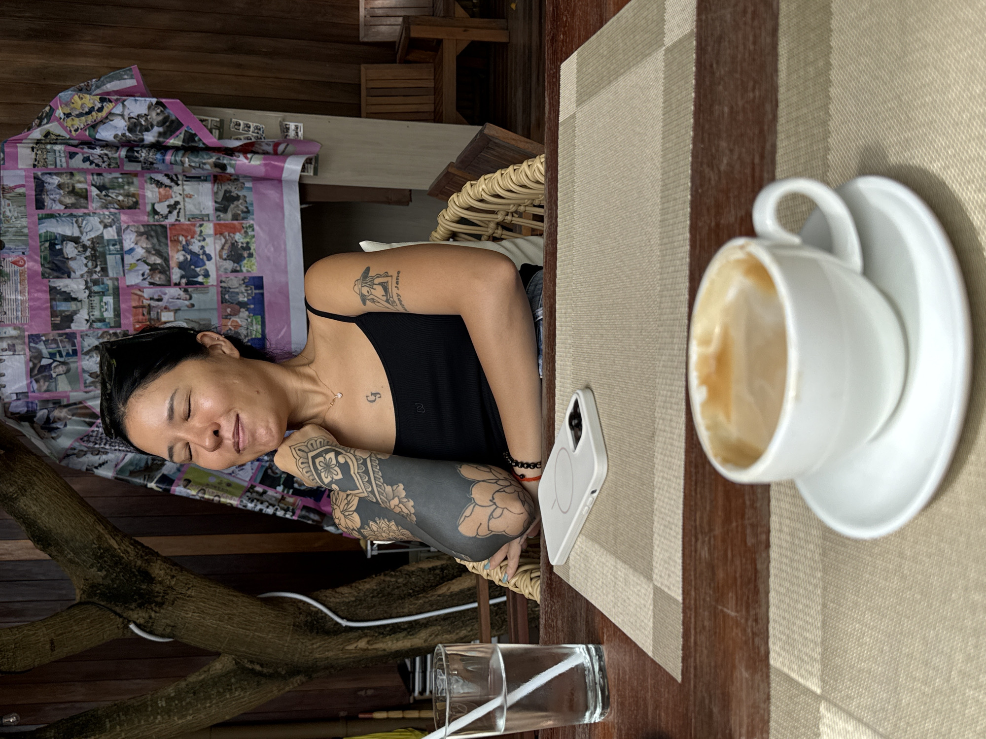 A medium shot captures a woman sitting at a table in what appears to be an outdoor cafe in Sai Ma Subdistrict, Thailand. The woman, with her eyes closed and a content expression, rests her chin on her hand. She has extensive black tattoos on her left arm and a smaller tattoo on her chest, along with a necklace. In front of her is a glass of water with a straw, a white mobile phone, and a cup of coffee on a saucer. The background features a wooden wall adorned with a collage of photographs, a tree trunk, and wooden chairs. The overall ambiance suggests a relaxed and casual setting.
