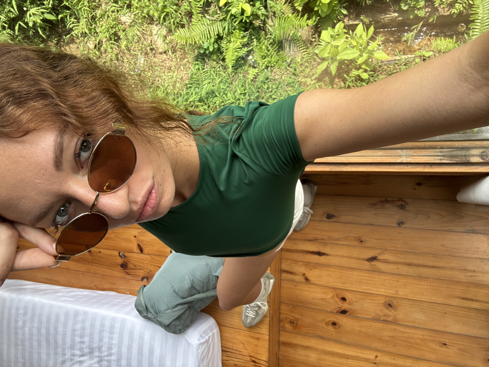 A young woman is captured in a high-angle selfie, looking up towards the camera with a slight head tilt. She has reddish-brown, wavy hair and wears amber-tinted sunglasses with gold frames, a dark green short-sleeved top, and light-colored shorts with matching sneakers. A light green or grey piece of fabric or clothing is draped around her left hip.
The setting appears to be a rustic indoor space, possibly a room in Du Già Commune, Vietnam, given the distinct wooden floorboards that cover the ground. To her left, part of a bed with white, striped bedding is visible.
Beyond a window or opening to her right, lush, vibrant green foliage, including ferns and other leafy plants, creates a natural backdrop. A hint of shallow water or a stream can be seen amidst the outdoor greenery, suggesting a serene natural environment.
The bright daylight indicates a daytime shot, illuminating the scene and highlighting the textures of the wood and the surrounding nature. There is no visible text in the image.