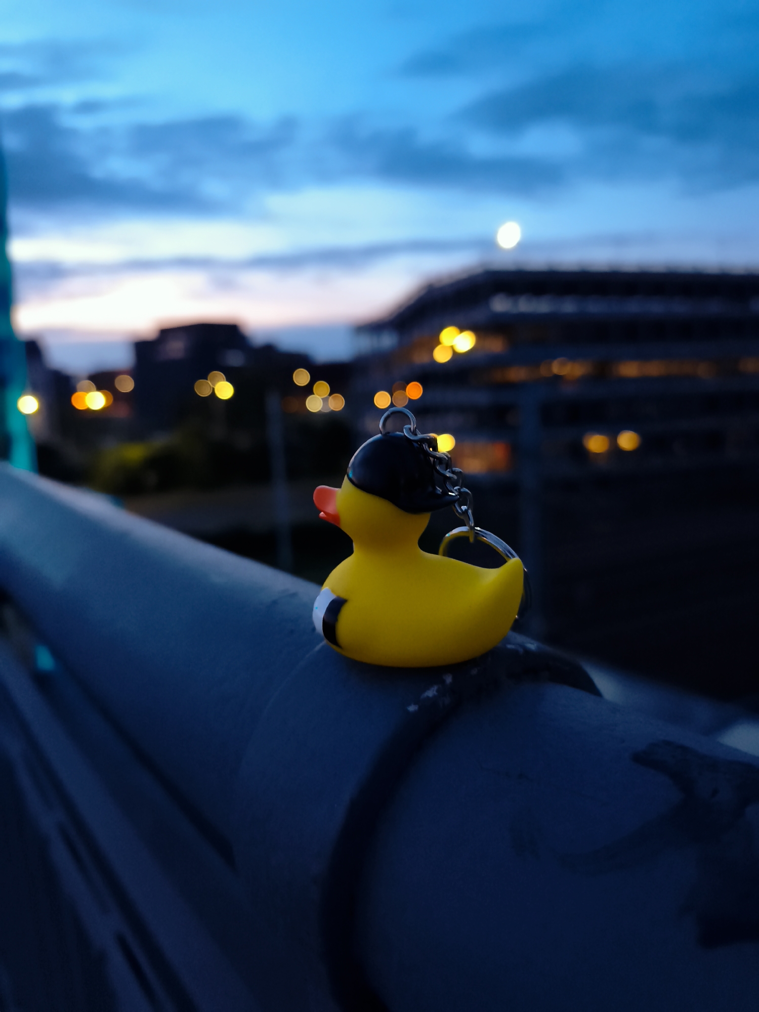 A yellow rubber duck keychain with a black head and orange beak is perched on a dark blue, textured railing. The duck faces to the right, with its head turned slightly towards the viewer. A silver chain connects to the duck's head and hangs down. The background is blurred, depicting a city skyline at dusk or night. Buildings with illuminated windows are visible, along with a faint moon in the deep blue sky. The overall scene suggests an urban environment. The location is identified as Gdańsk, Poland.