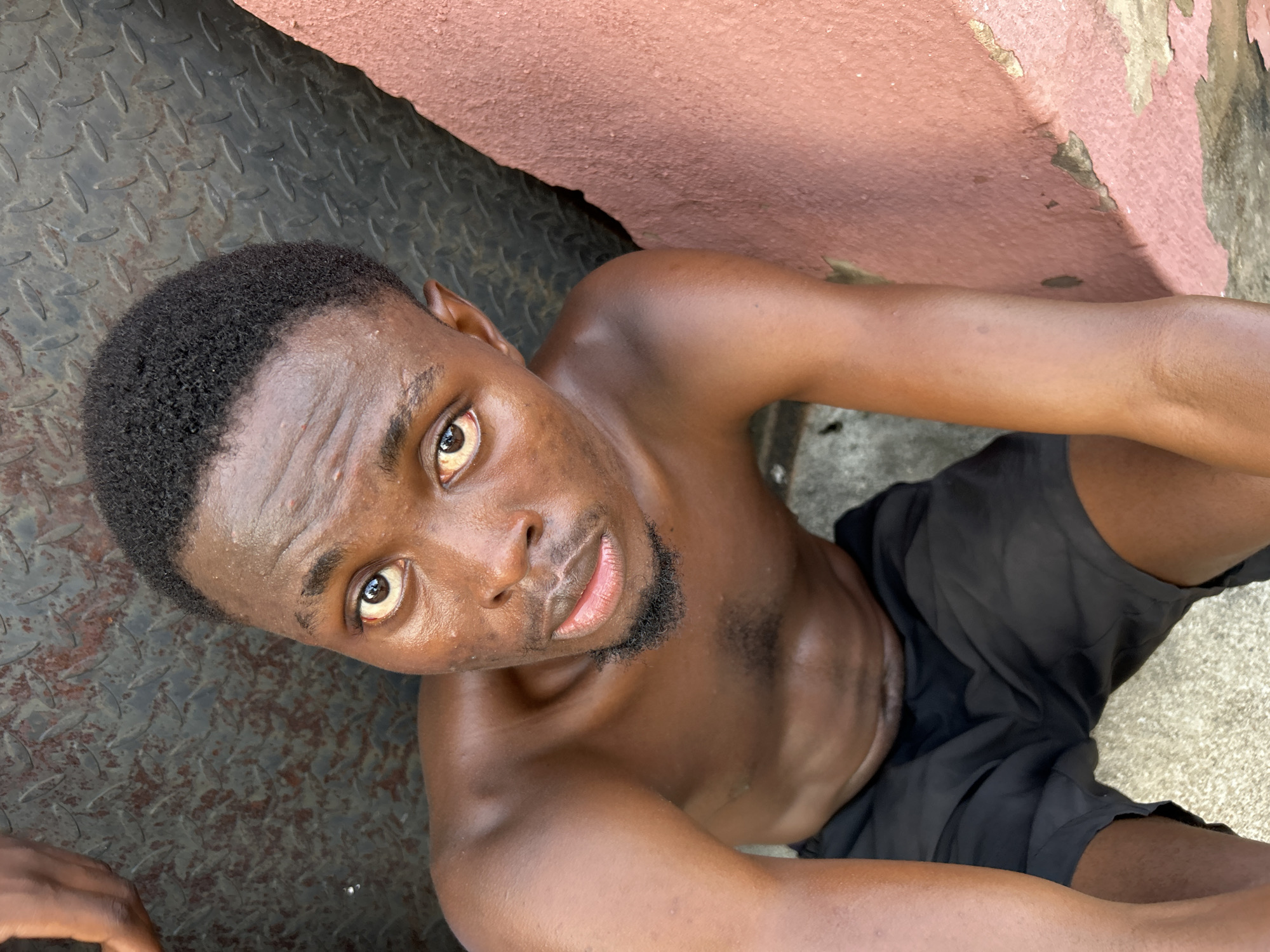A close-up, high-angle shot depicts a shirtless, dark-skinned man with a short beard and close-cropped hair. He is looking up into the camera. He is wearing black shorts. The background includes a mottled gray metal surface on the left, a pink painted wall above, and a concrete area below. The location is in Ihiagwa, Nigeria.