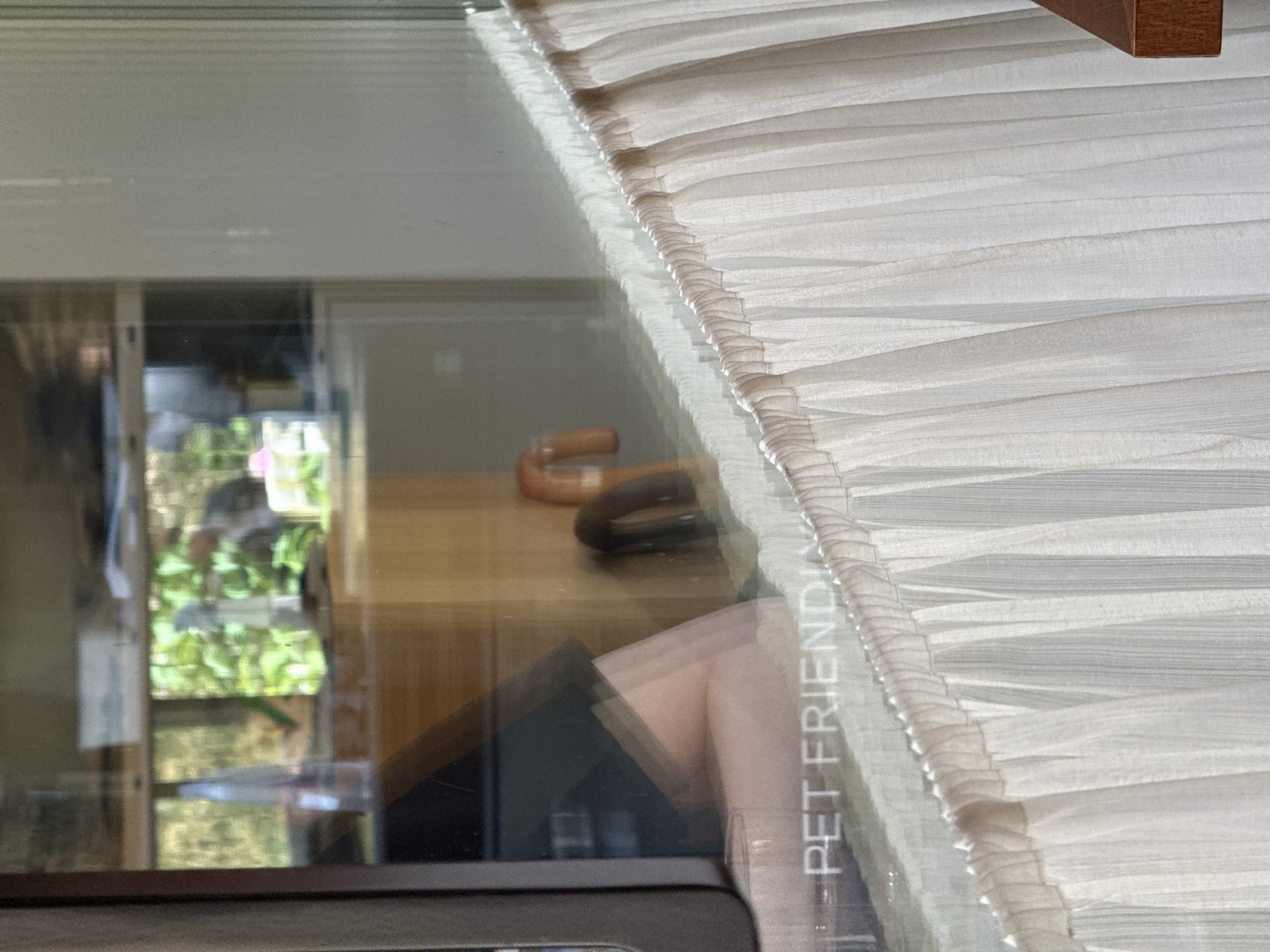 The image shows a partial reflection in a glass surface. The reflection includes an umbrella and a book with the words "PET FRIENDLY" printed on it. The environment reflected is a room interior. There is greenery visible through a window, suggesting an outdoor area. The image also includes a white, layered fabric in the foreground and an out-of-focus wooden object in the top right corner. The city and location of Belgrade, Serbia cannot be determined from the image.