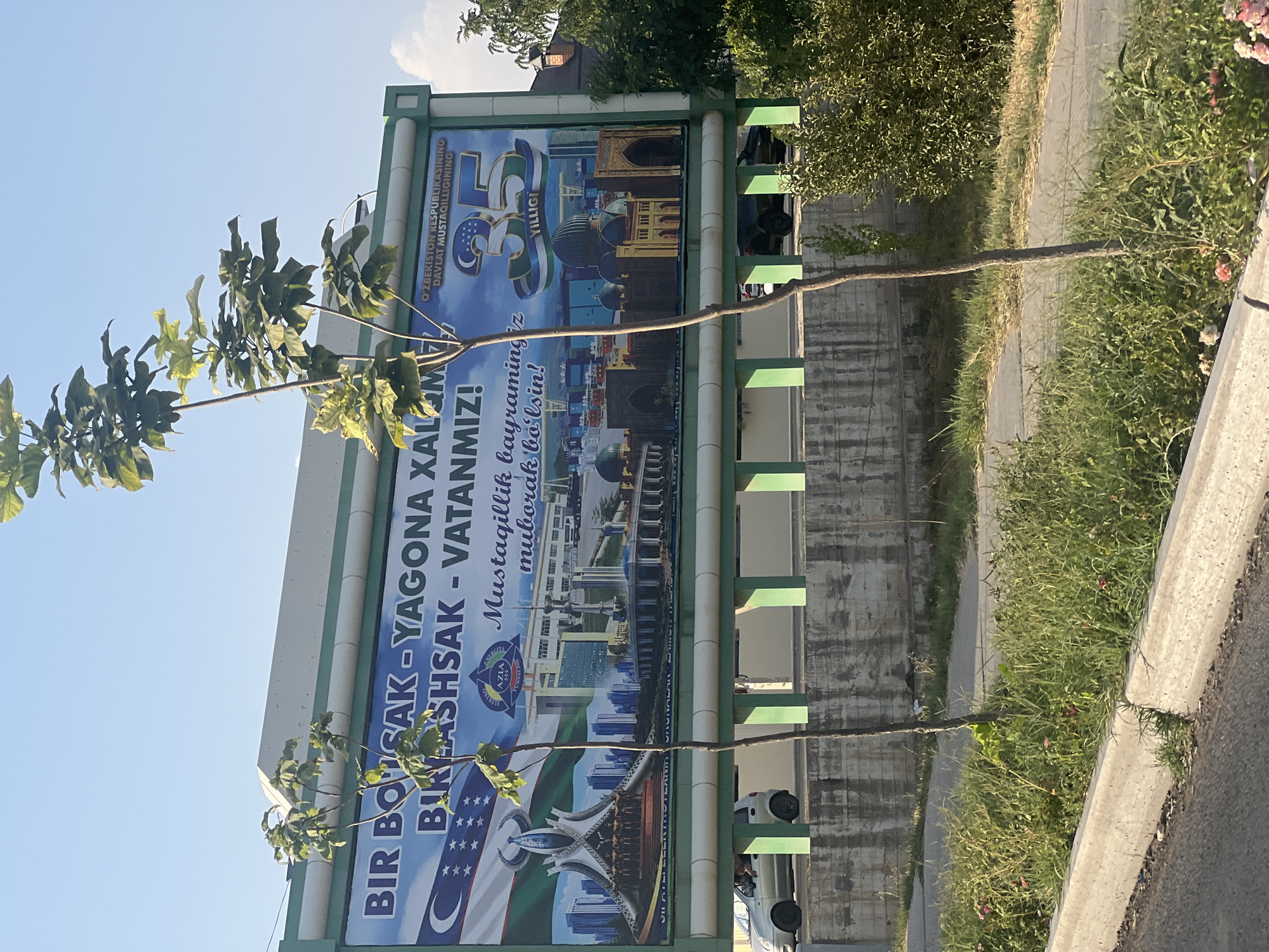 The image captures a billboard situated on the side of a road in Kibray, Uzbekistan, on a clear, sunny day. The billboard, a large rectangular structure, is predominantly blue and features text in Cyrillic and Latin scripts, along with images of Uzbek national symbols and landmarks. The text appears to be a celebratory message, possibly related to a national holiday, with phrases like "Mustaqillik bayramingiz muborak bolsin!" which translates to "Happy Independence Day!". The billboard also displays a prominent "35" with the Uzbek flag superimposed, suggesting it commemorates the 35th anniversary of something significant.
The billboard is framed by a green metal structure. In front of the billboard, two young trees with sparse foliage are growing, their thin trunks and branches extending upwards. The foreground shows a grassy area with some small pink flowers and a concrete curb bordering a paved pathway. Beyond the billboard, a concrete wall is visible, with cars driving on a road behind it. The overall impression is one of public commemoration and civic pride in a seemingly suburban or semi-urban environment in Uzbekistan.