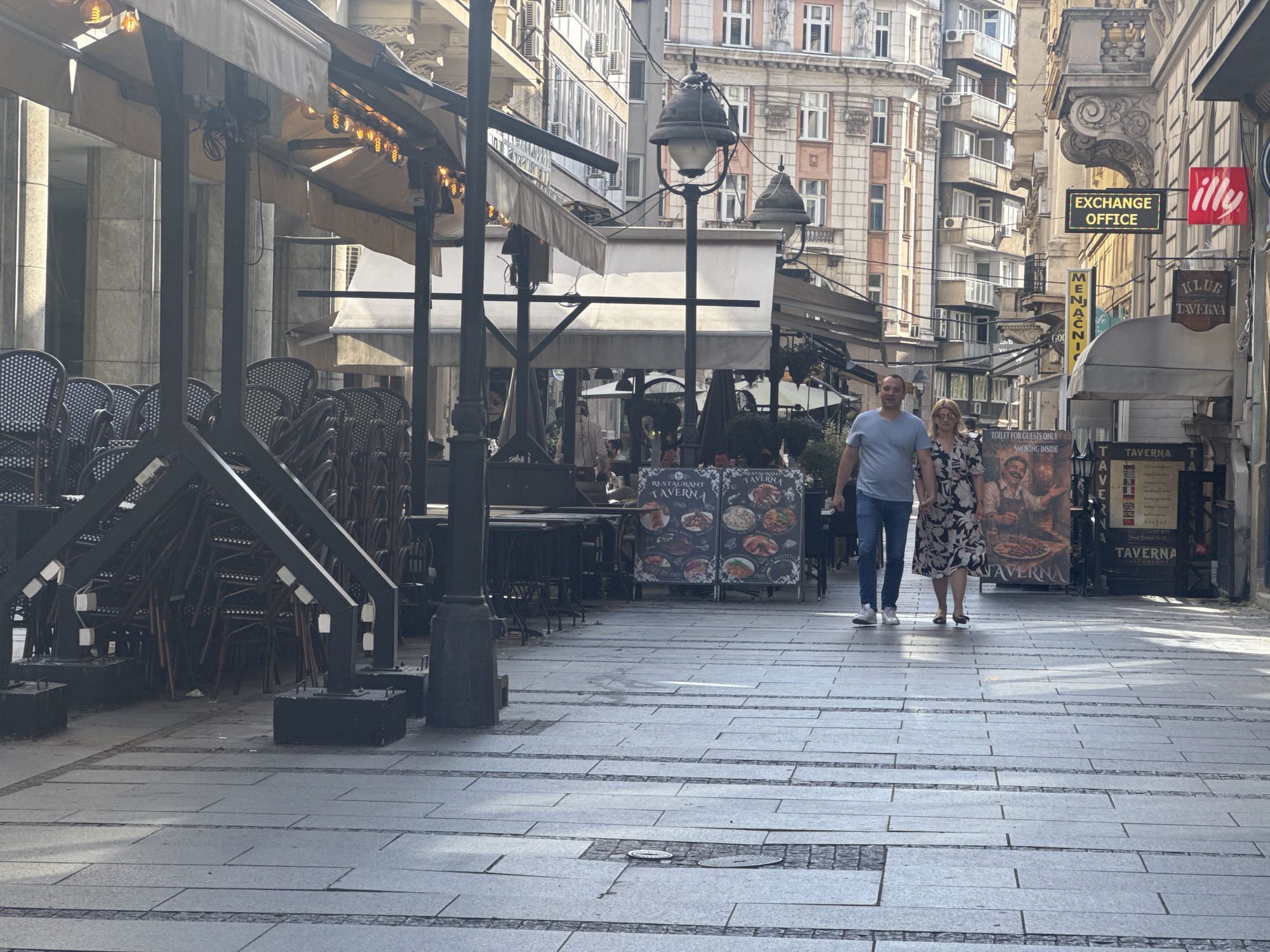 This image depicts a street scene in Belgrade, Serbia, likely Kalemegdan Fortress or a pedestrian street in the city center, given the architectural style of the buildings and the presence of outdoor cafes.
In the foreground, a man and a woman are walking towards the viewer on a paved pedestrian walkway. The man is wearing a light blue t-shirt and blue jeans. The woman is wearing a floral print dress. They are holding hands and appear to be casually strolling.
To the left of the walkway, several outdoor cafe seating areas are visible. Chairs are stacked and covered with awnings. Large signs advertising food and drinks for "RESTAURANT TAVERNA" are displayed.
The background features a street lined with buildings showcasing a mix of architectural styles, typical of Belgrade. Some buildings have classical facades with ornate details, while others appear more modern with balconies and air conditioning units. Signs for businesses such as "EXCHANGE OFFICE" and "illy" are mounted on the buildings. The scene is bathed in sunlight, suggesting it is daytime.