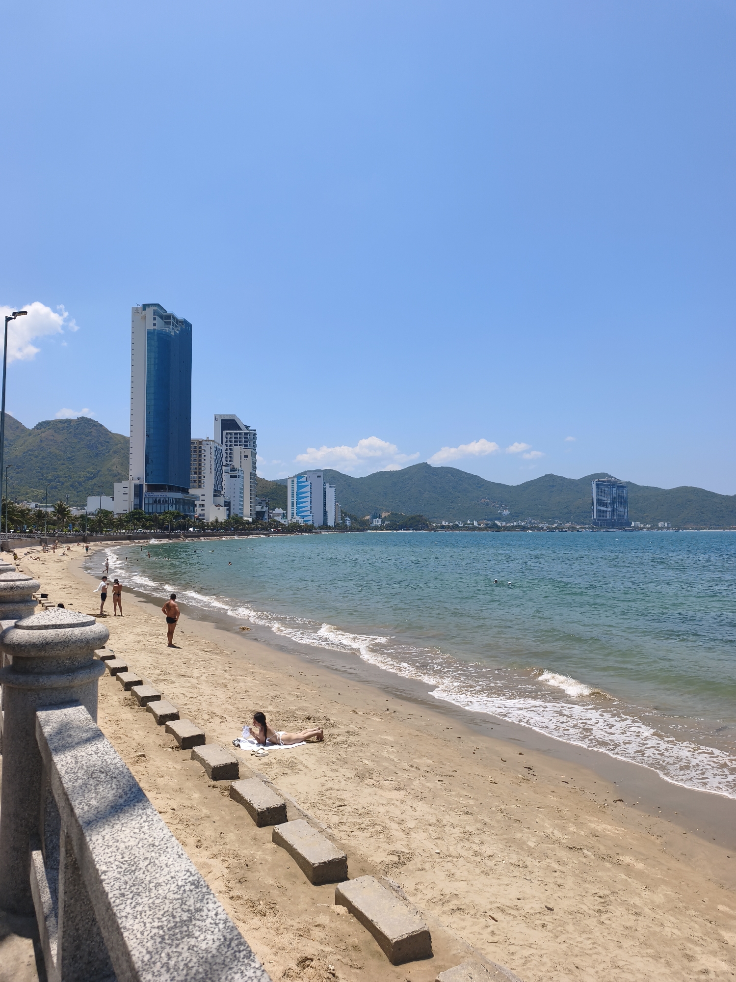 The image depicts a view of Nha Trang beach in Vietnam on a clear, sunny day. The foreground shows a textured stone railing and a sandy beach with several concrete blocks arranged in a line. A woman in a white bikini is lying on a towel on the sand, facing away from the camera. Further up the beach, two people are standing near the water's edge, one in dark swim trunks and the other in a black bikini. In the background, the turquoise ocean meets the sandy shore, with gentle waves lapping the beach. A line of palm trees and other foliage separates the beach from a street, behind which rise several modern high-rise buildings, including a prominent blue-glass skyscraper. Beyond the buildings, lush green mountains form a backdrop under a clear blue sky with a few wispy white clouds. The scene conveys a typical beach resort atmosphere with a blend of natural beauty and urban development.