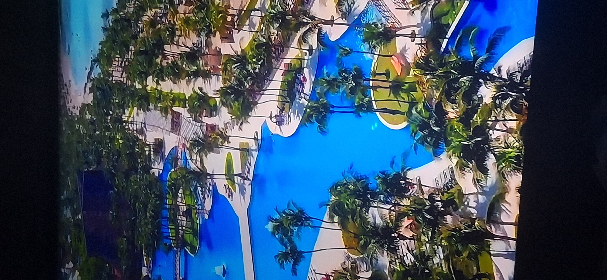 The image shows an aerial view of a resort with multiple swimming pools, surrounded by palm trees and a beach in the background. The pools are a vibrant blue and have winding shapes with white pathways connecting different sections. There are lounge chairs and umbrellas placed around the pool areas. A small white boat is visible in one of the pools. The resort appears to be located in a tropical or subtropical environment, with lush green vegetation. There is no information in the image to suggest it is located in Bila Tserkva, Ukraine.