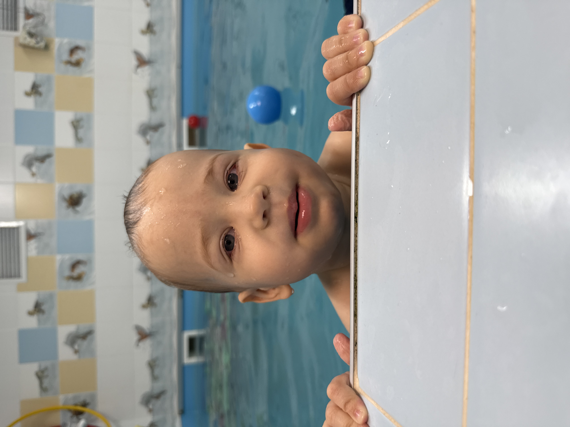 A child with wet hair is clinging to the tiled edge of a swimming pool. A blue ball floats in the water behind the child. The wall in the background is tiled with a pattern of alternating blue and yellow squares, each containing a depiction of birds. The location is presumed to be Rusanovo, Russia, based on the prompt.
