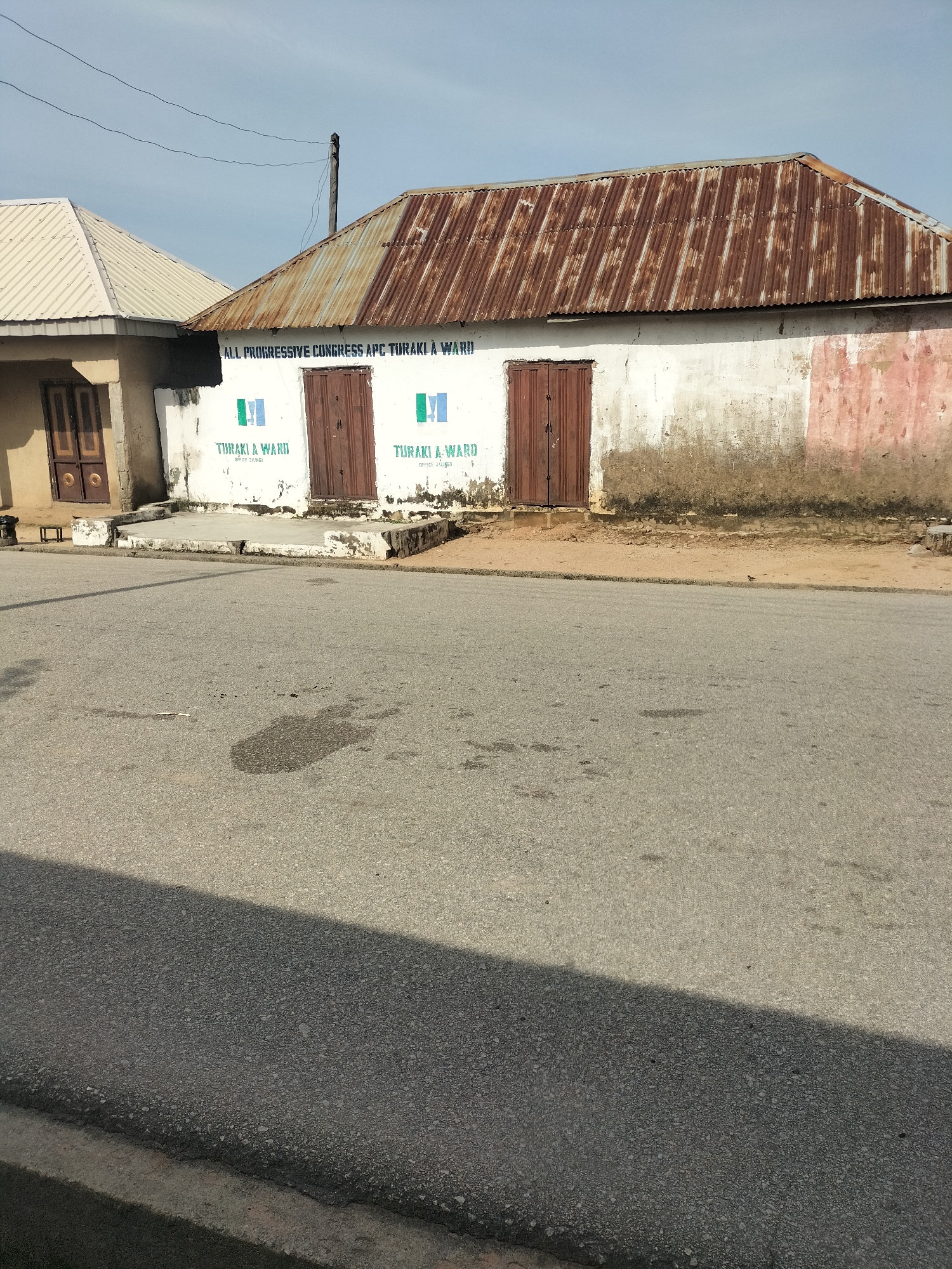 The image captures a weathered building in Jalingo, Nigeria, with a corrugated iron roof that is heavily rusted and shows signs of age. The building's exterior walls are a pale white, with patches of peeling paint and dirt, particularly near the base. Two brown wooden doors are visible on the front of the building. Above the doors, text is painted on the wall, reading "ALL PROGRESSIVE CONGRESS APC TURAKI A WARD" and "TURAKI A WARD OFFICE JALINGO" with the APC party logo. To the left, another building with a silver corrugated iron roof is partially visible, featuring a brown door and a light-colored wall. The foreground consists of an asphalt road, with some dark stains on its surface. The sky above is clear and light blue. The overall impression is one of an older structure in a somewhat neglected state, located in a community setting.