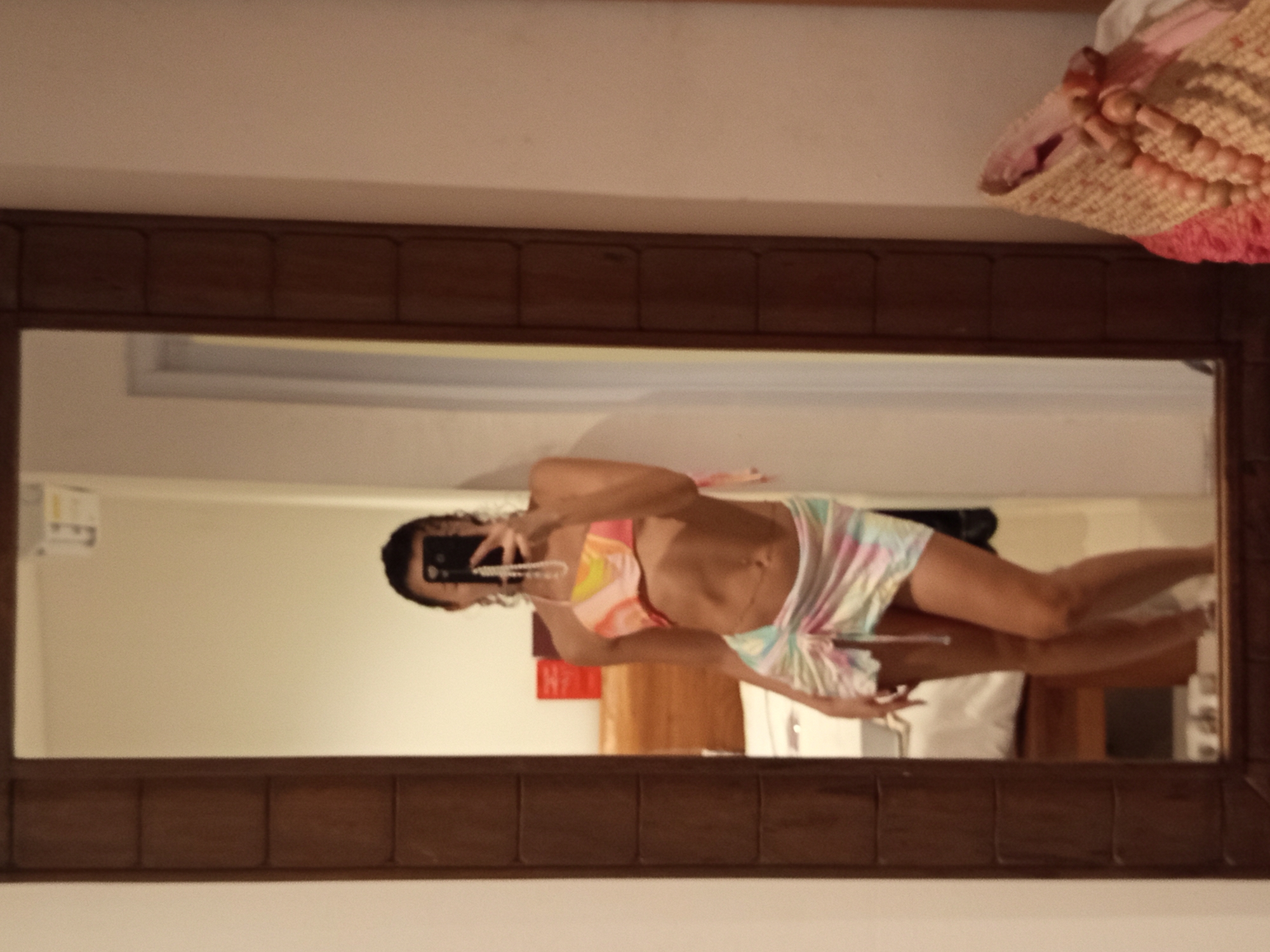 A woman is taking a mirror selfie indoors. The mirror has a dark wooden frame. The woman is wearing a colorful bikini top and a matching skirt. There is a partially visible straw bag with pink accents and wooden beads on top of the mirror's frame. The room appears to be a hotel room based on the furniture and decor visible in the mirror. It is plausible that this image was taken in North Kuta, Indonesia, as that is part of the prompt, but this cannot be confirmed definitively from the image alone.