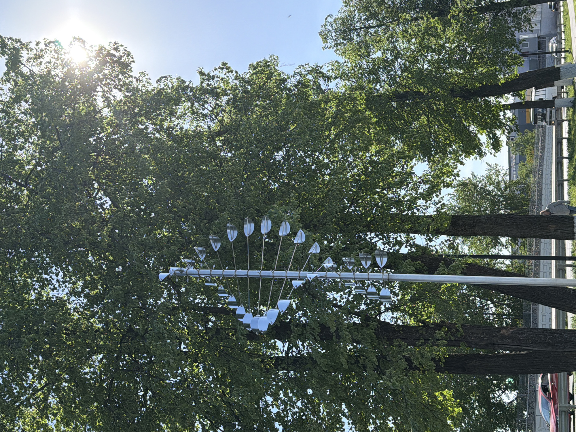 The image shows a street in Votkinsk, Russia, under a sunny sky. A contemporary art installation featuring reflective, spoon-shaped elements mounted on a horizontal metal bar is positioned among trees, creating a contrast between the natural foliage and the artificial structure. Trees line the street. The background includes buildings and parked vehicles, indicative of an urban environment.