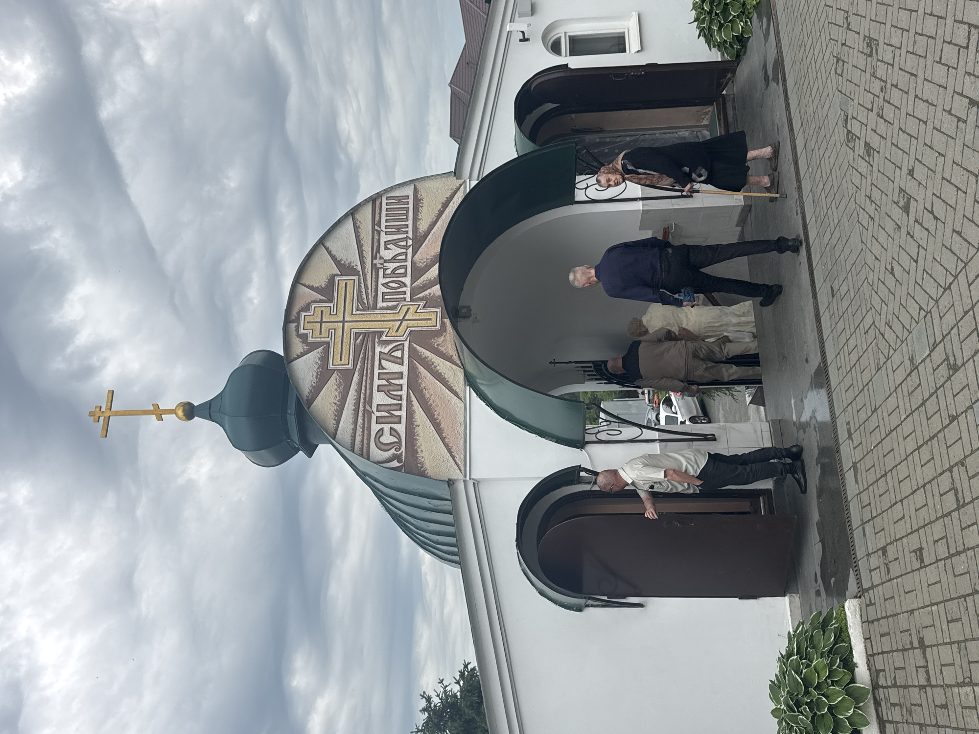 The image shows the entrance to a white building with a green domed roof topped by a golden cross. Above the arched entrance, a circular mosaic features a golden Orthodox cross and what appears to be text in Cyrillic. Several people are visible: a man in a light-colored shirt and dark pants stands at an open doorway on the left, holding it open. Inside the entrance, a group of people are seen walking up steps. To the right of the entrance, a woman in a black dress and headscarf leans on a walking stick, facing the building. The ground in front of the building is paved with interlocking gray bricks, and there are plants on either side of the entrance. The sky is overcast with dramatic clouds. The building appears to be the Church of the Icon of the Mother of God "Joy of All Who Sorrow" located on Malysheva Street in Yekaterinburg, Russia.
