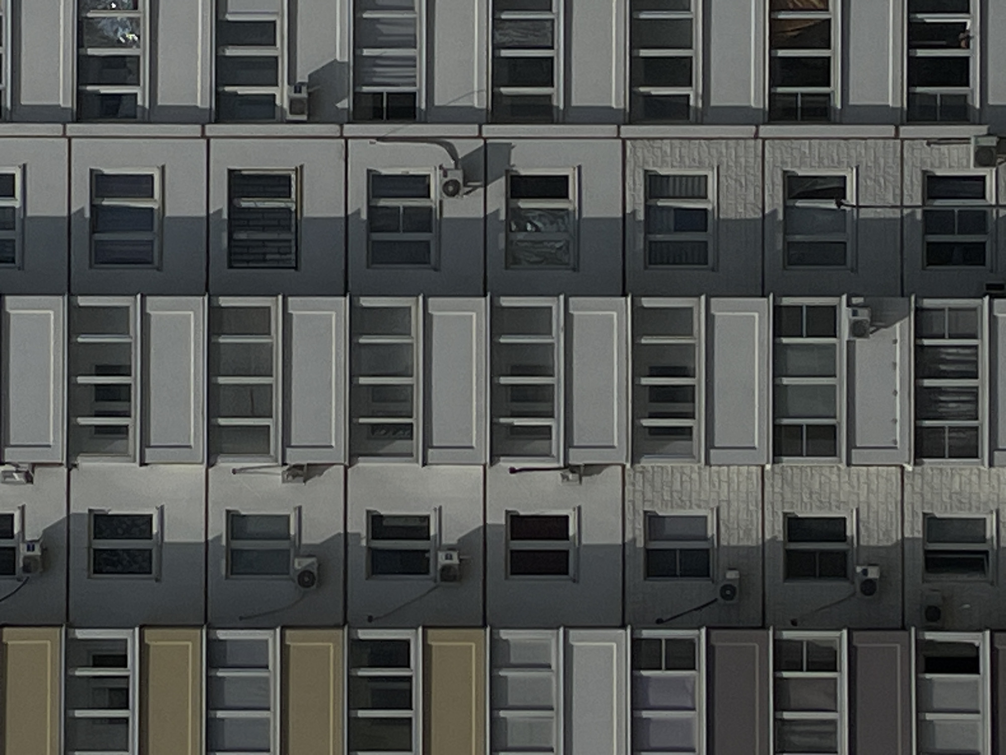 The image depicts the facade of a Soviet-era apartment building, likely in Rostov-on-Don, Russia, characterized by its uniform structure and repetitive architectural elements. The building is constructed with precast concrete panels, typical of mass housing projects from that era, and is painted in a muted, light gray color.
The facade is divided vertically into three main sections, with each section containing multiple floors. Each floor is further subdivided into individual apartments, each featuring a balcony and windows. The balconies are enclosed with railings, some of which appear to be made of metal.
Windows are a dominant feature, appearing in various configurations on each apartment. Many windows are large and rectangular, with white frames. Some windows are open, revealing the interiors, while others are closed. The balconies have a consistent design, with a light gray border and a darker gray or yellowish color on the outer panels of some floors, suggesting different materials or finishes.
Several air conditioning units are mounted on the exterior walls below some of the windows, indicating a modern upgrade to the older building. The lighting suggests it is daytime, with harsh shadows cast by the architectural features, implying sunlight. The overall impression is one of urban uniformity and functional architecture, common in post-Soviet cities. No people are visible in the image.